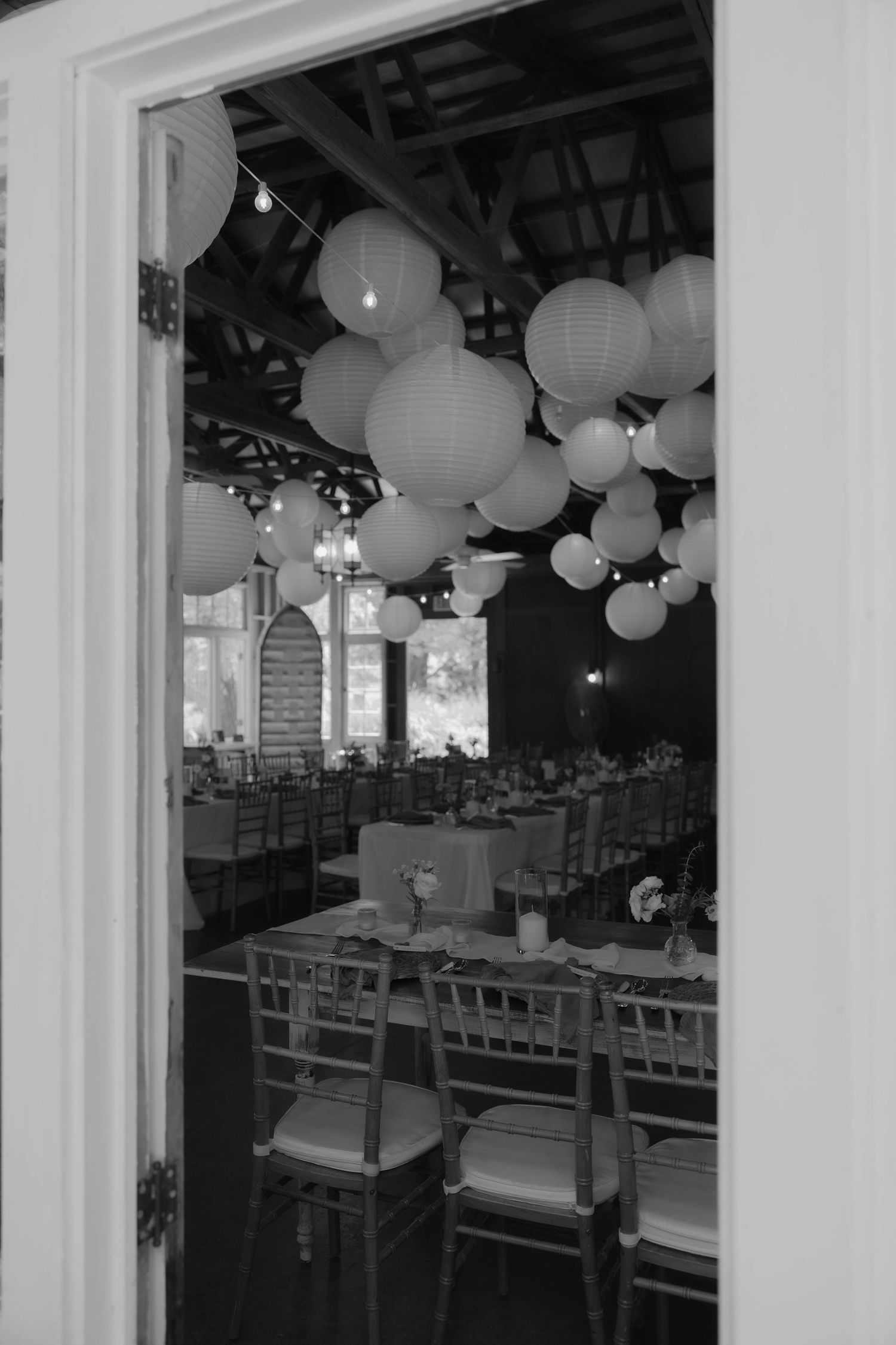 Black and white photo of elegant event hall with paper lantern ceiling decor, chiavari chairs, and long dining tables set for a reception.