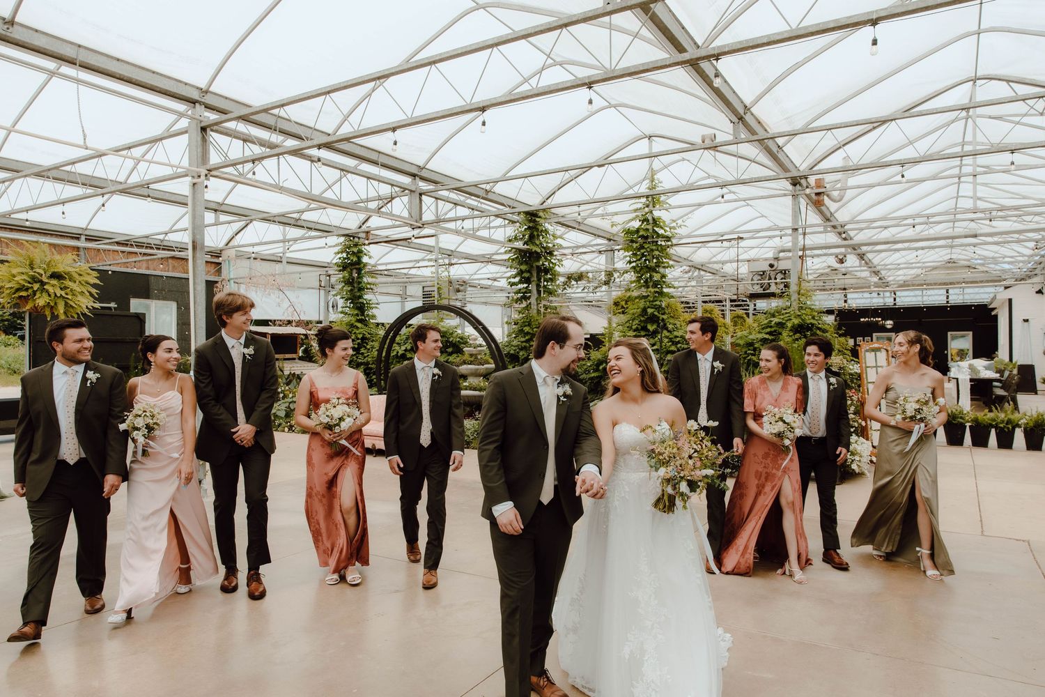 Wedding party walks through a greenhouse venue, bride and groom at center, bridesmaids in mixed earth tones.