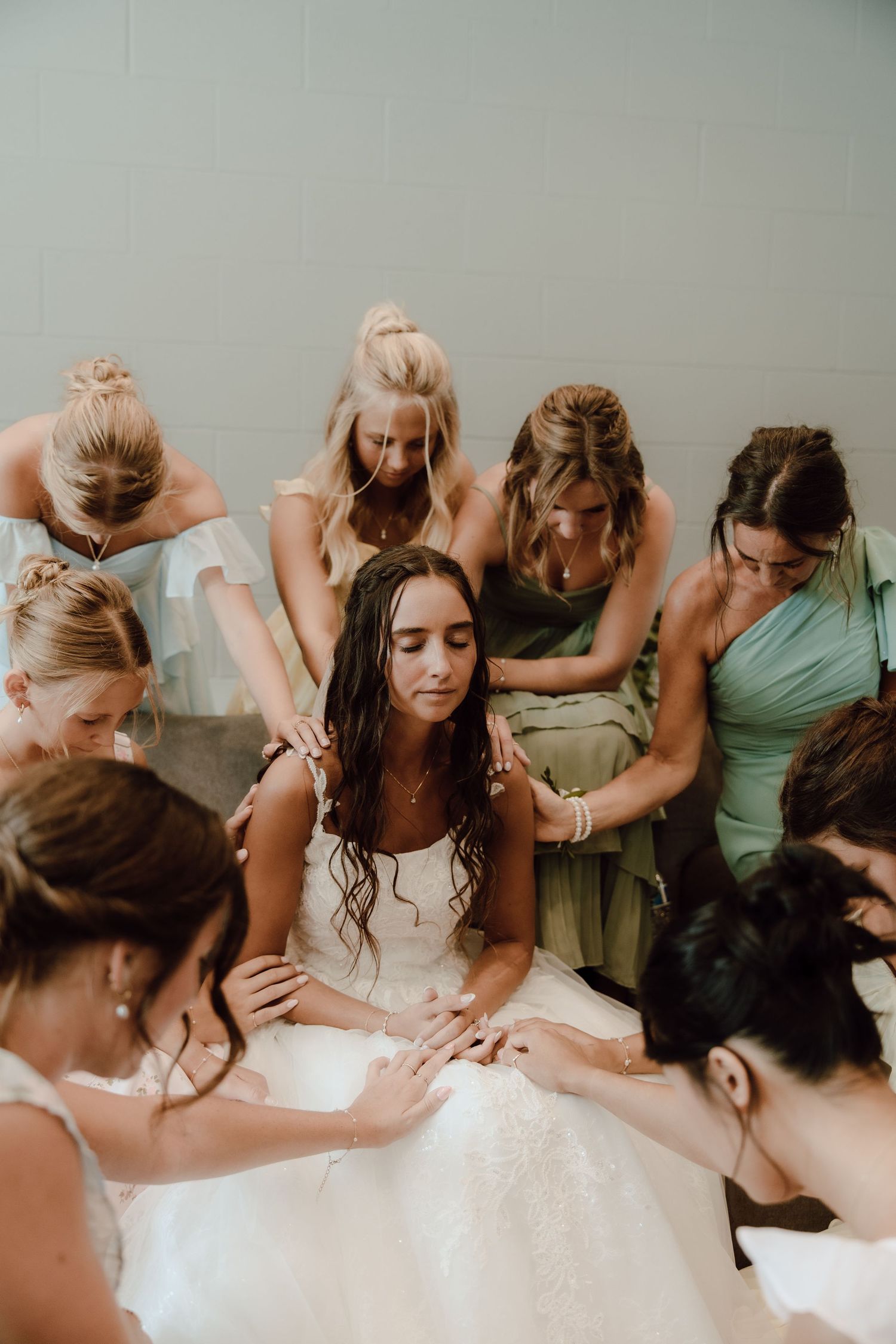 Bride in white gown surrounded by bridesmaids in green dresses praying together before wedding ceremony.