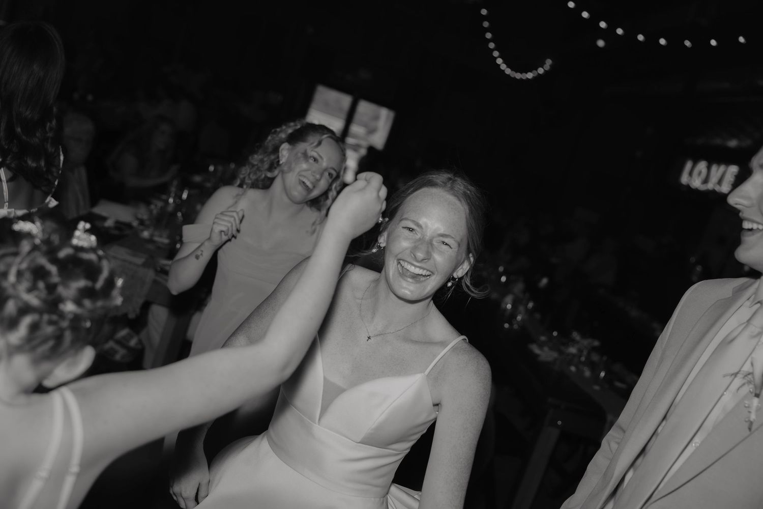 Black and white photo of a laughing bride in a white dress surrounded by smiling guests at a wedding reception.