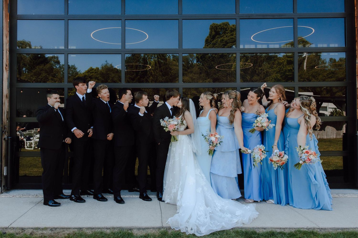 Wedding party poses outside modern venue, groomsmen in black suits and bridesmaids in blue gowns flanking bride and groom.