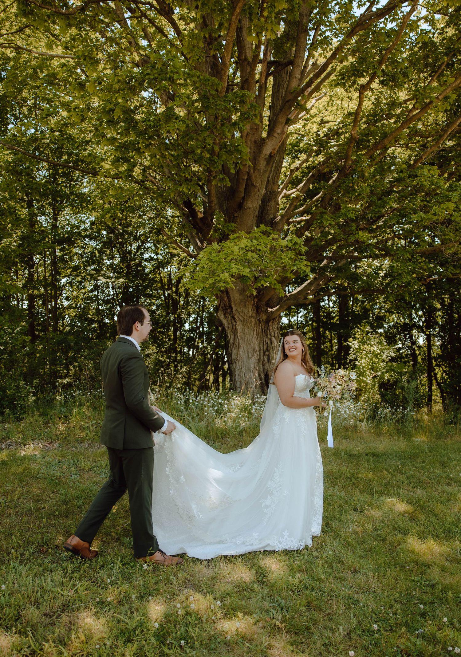 Bride in white gown holds bouquet as groom carries her train in a lush green outdoor wedding setting.