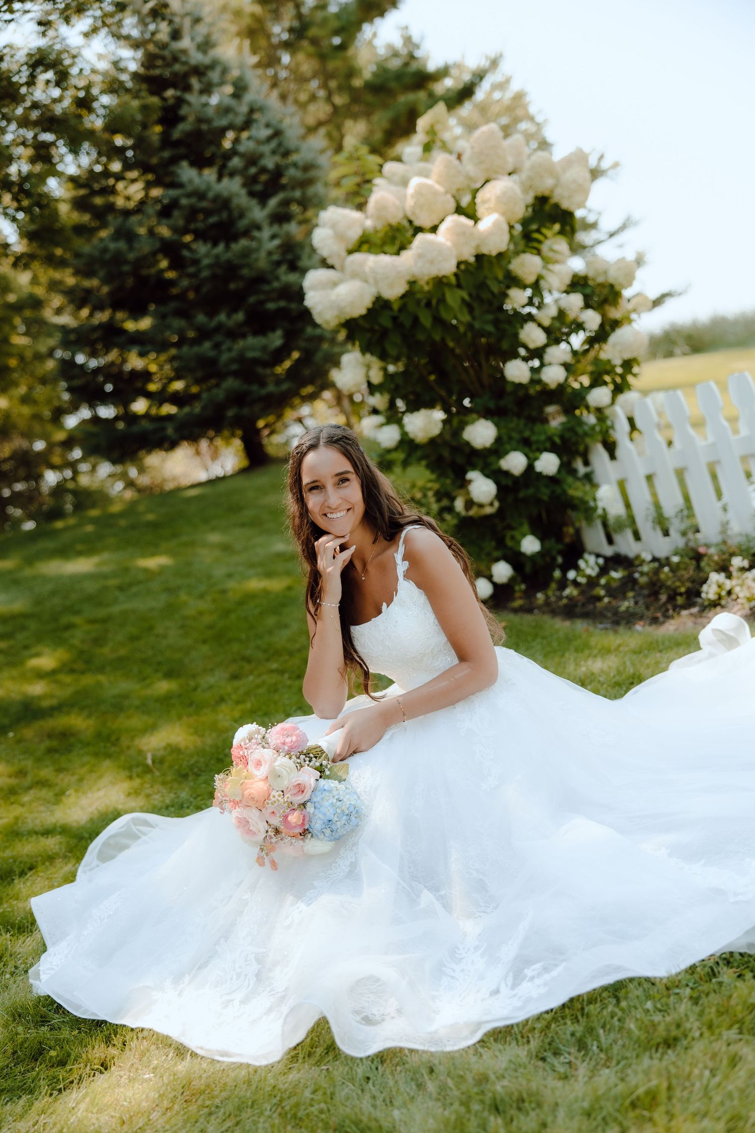 Smiling bride in white ball gown sits on grass holding pastel bouquet near white hydrangea bush.