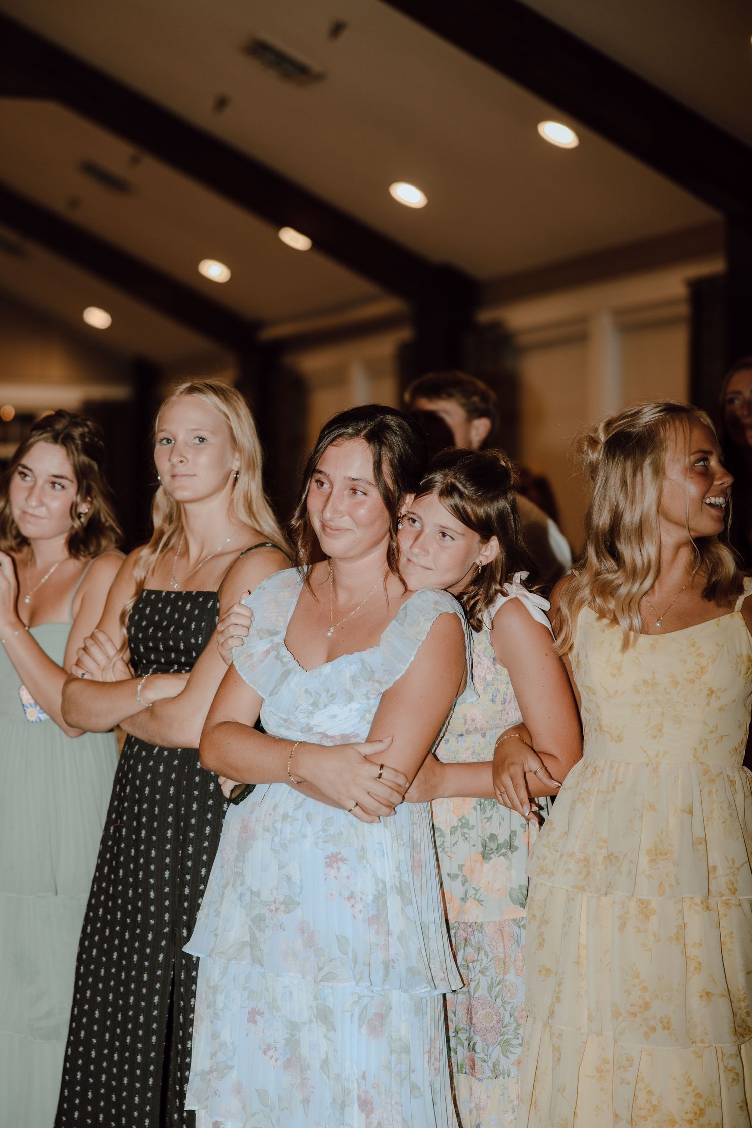 Group of young women in floral and patterned summer dresses standing together at an indoor wedding reception.