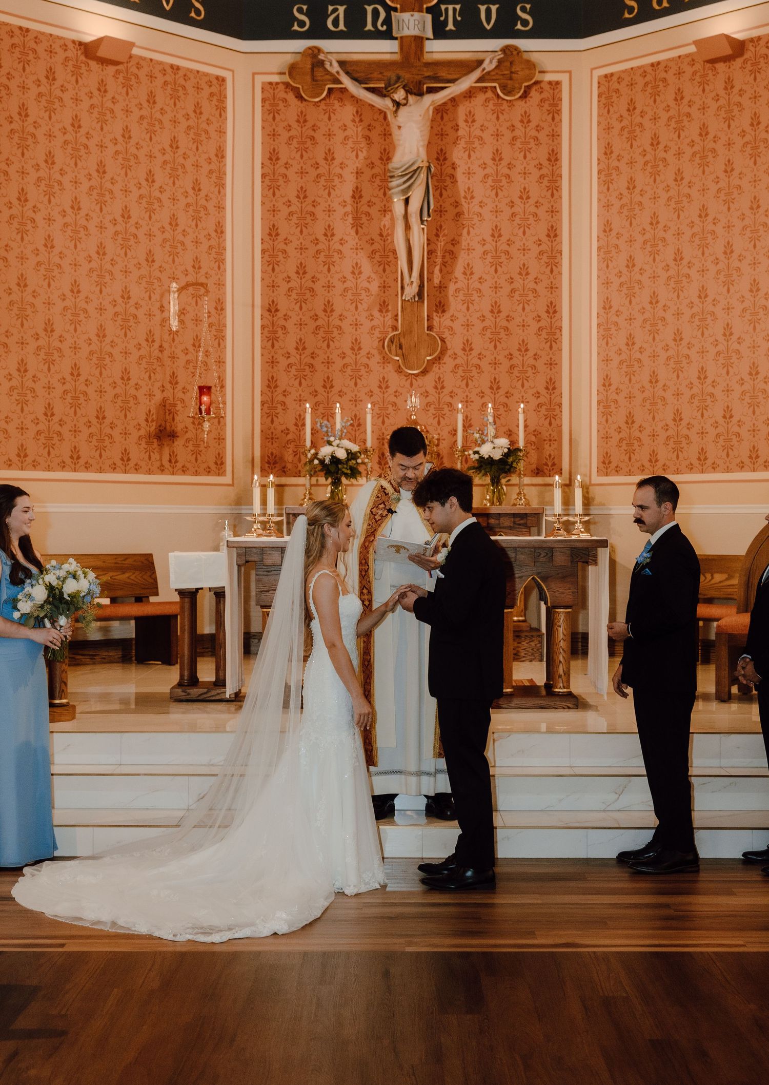 Bride and groom exchange vows at a Catholic church altar with crucifix, candles, and wedding party during ceremony.