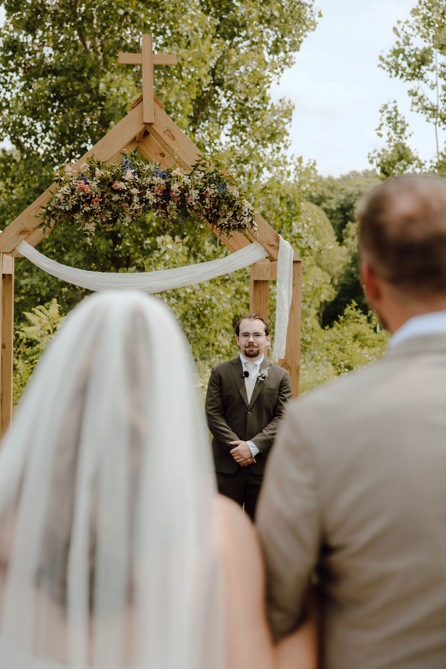 Groom stands at floral wooden arch altar as bride walks down the aisle with escort at outdoor wedding.