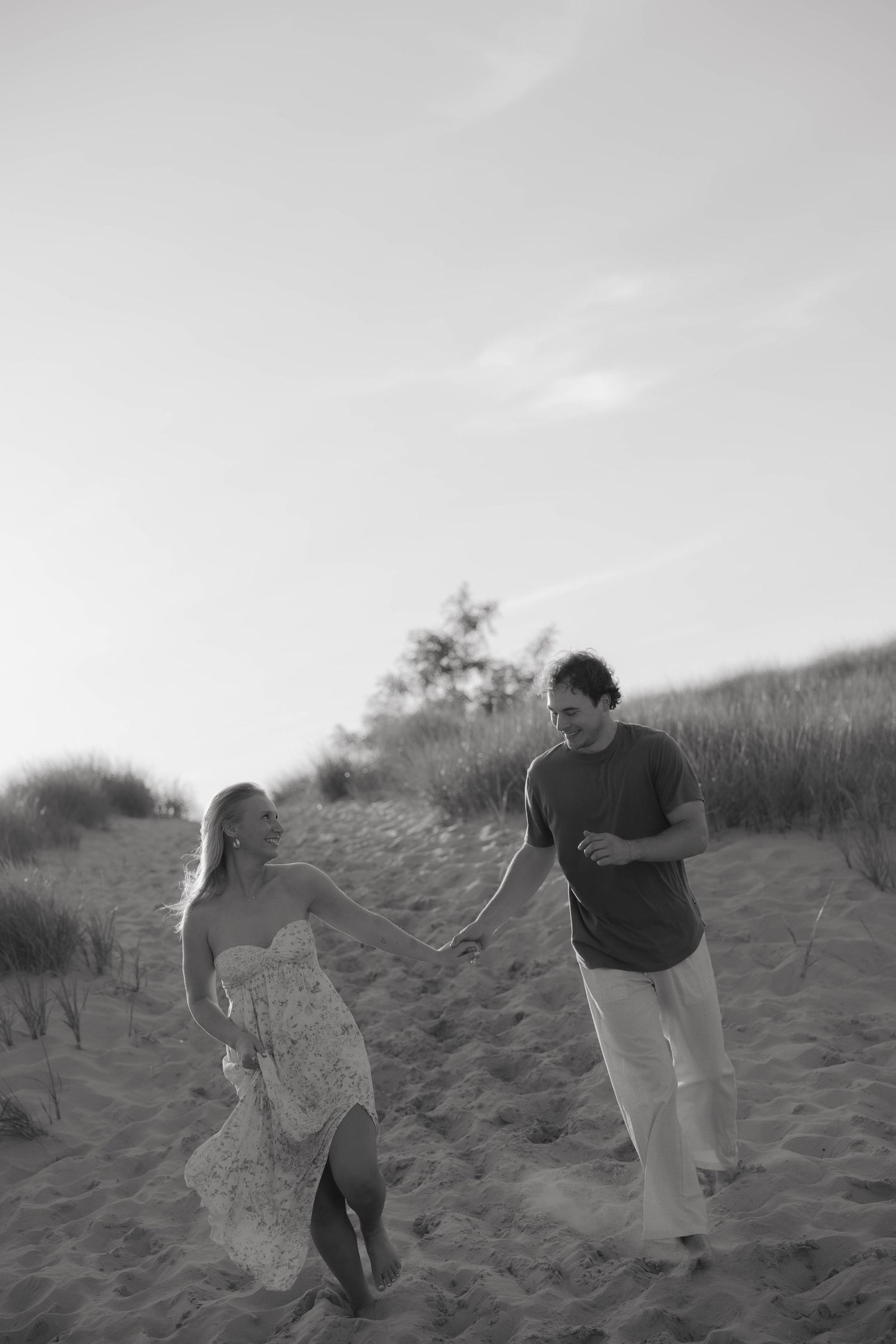 Black and white photo of a couple running and laughing on sandy beach dunes.