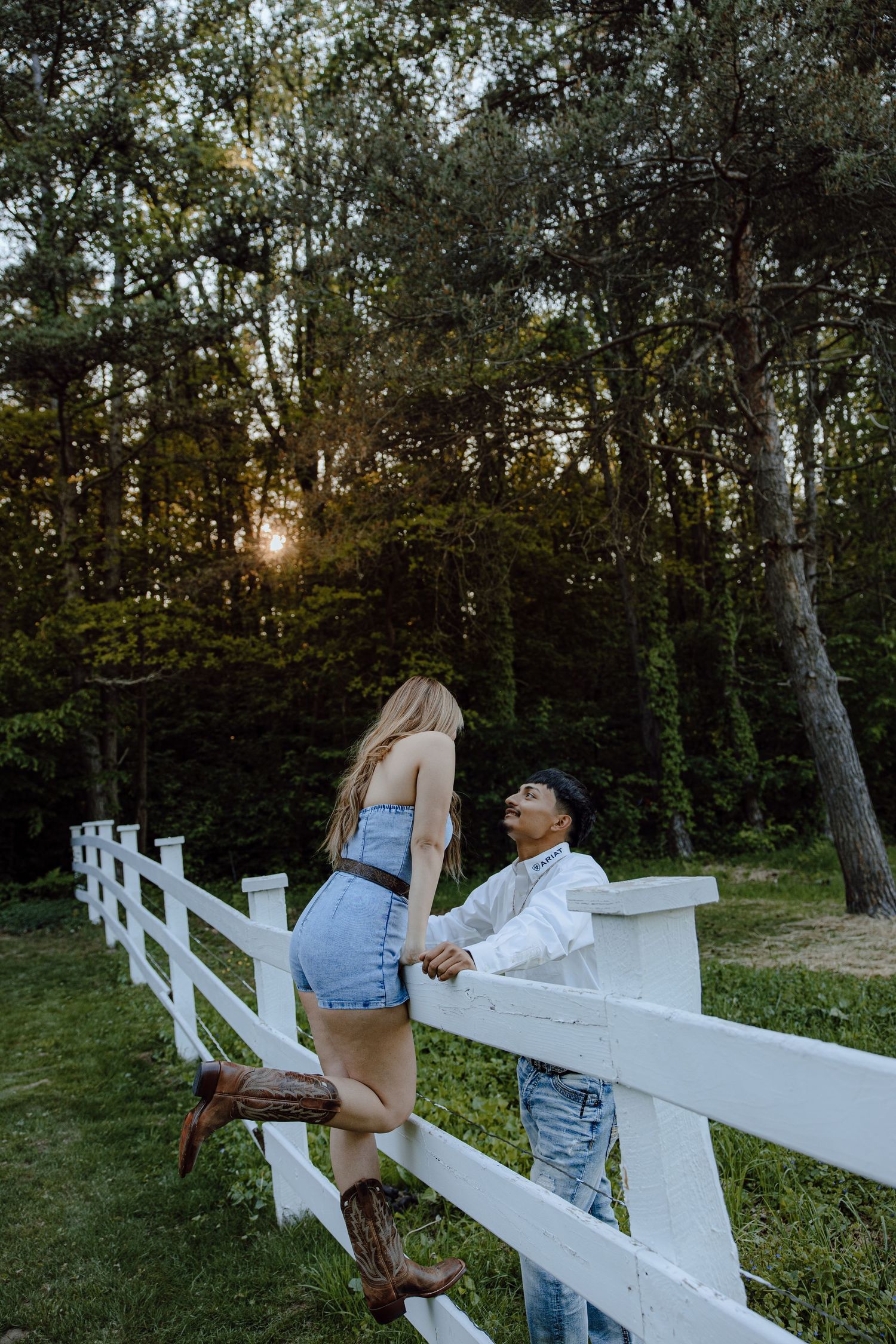 Playful couple interaction by white fence at sunset in wooded area.