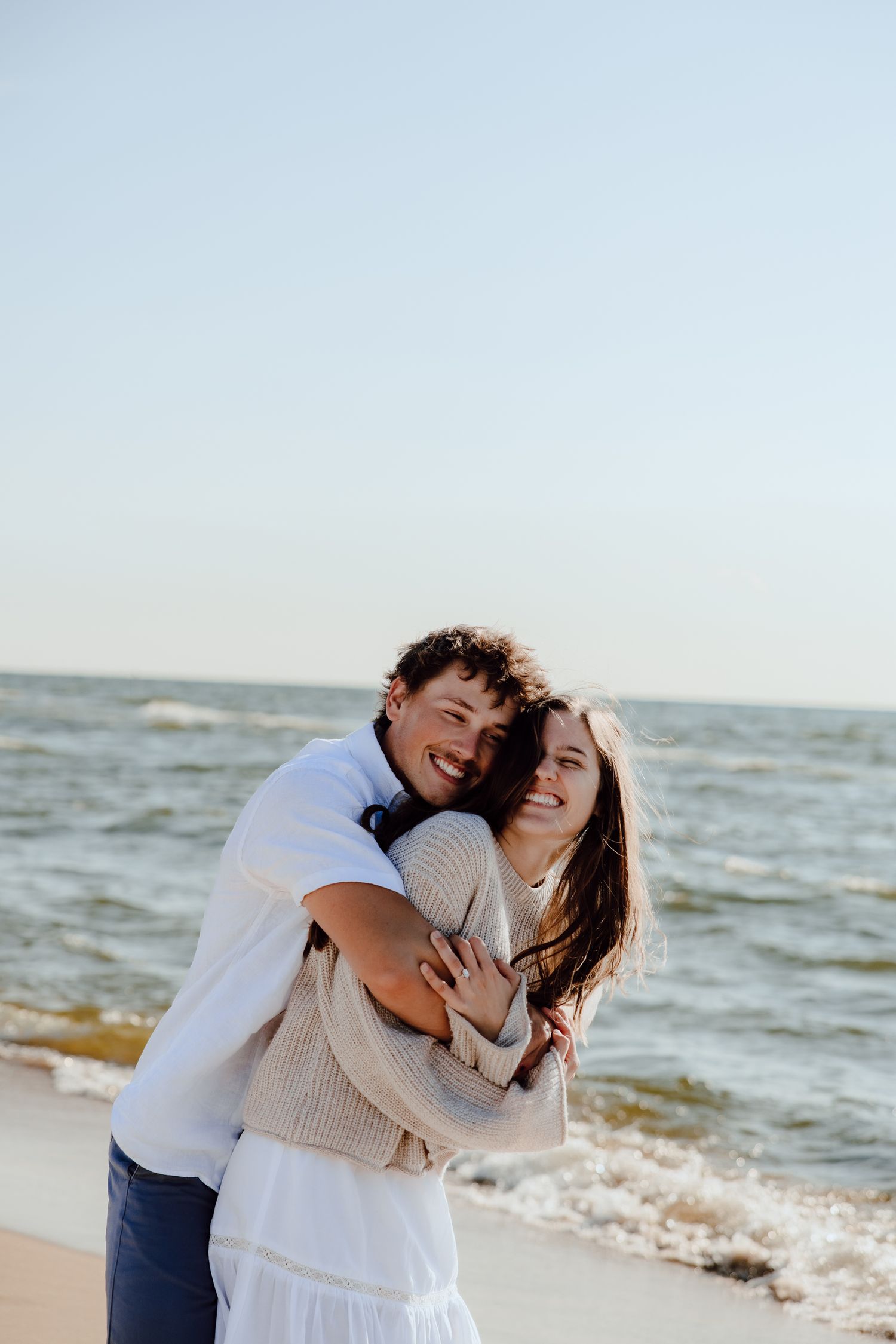 A couple embraces and laughs together on a sandy beach with ocean waves in the background.