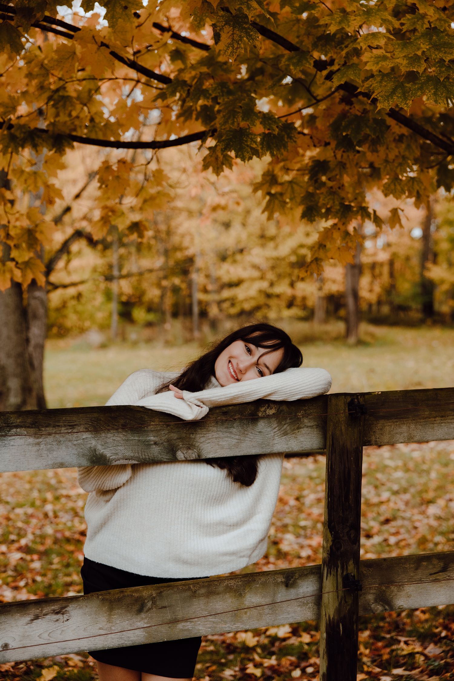 Person in cozy white sweater lounges on wooden fence in autumn park setting.