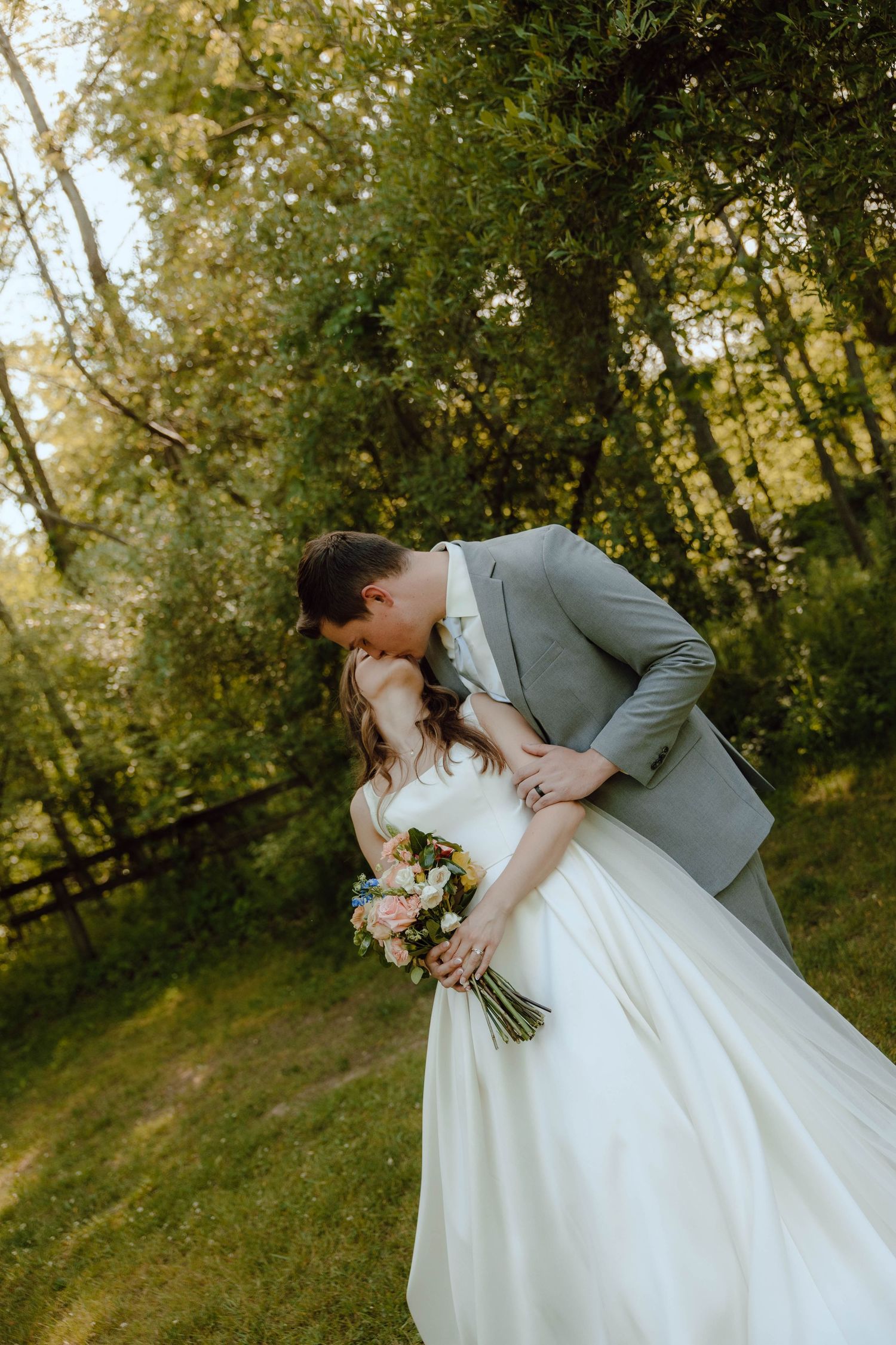 Groom in gray suit dips bride in white gown for a romantic kiss outdoors surrounded by lush green trees.