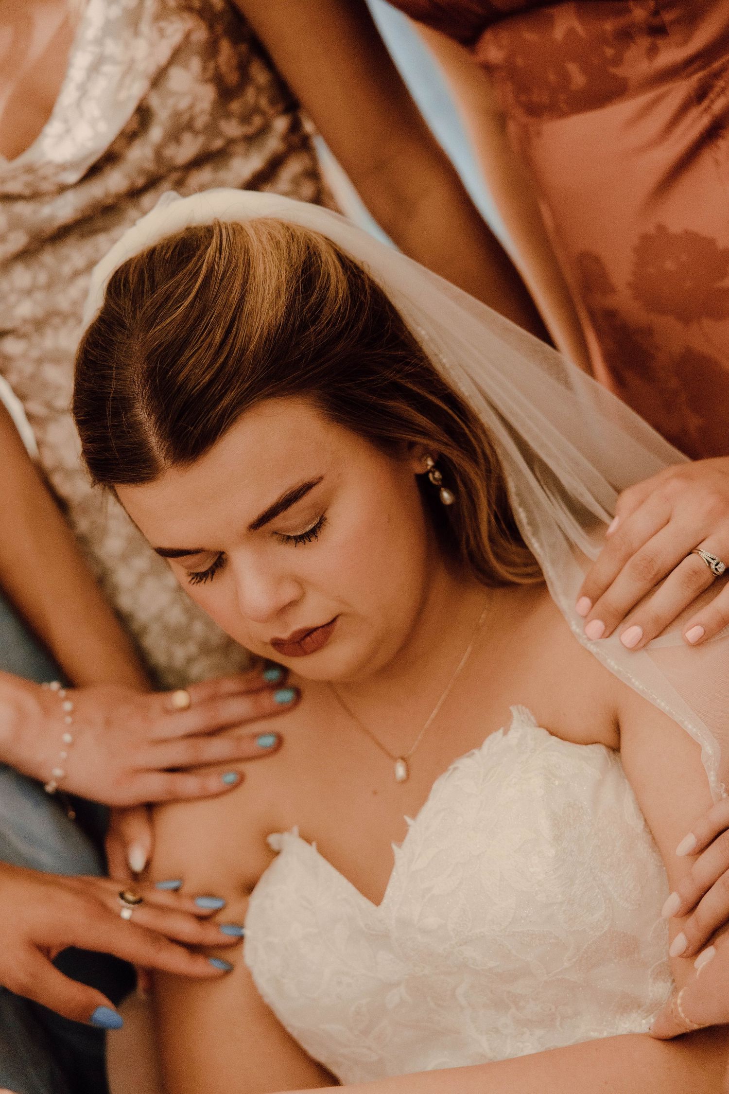 Bride in white lace strapless gown with veil being helped by bridesmaids in pink and gold dresses.
