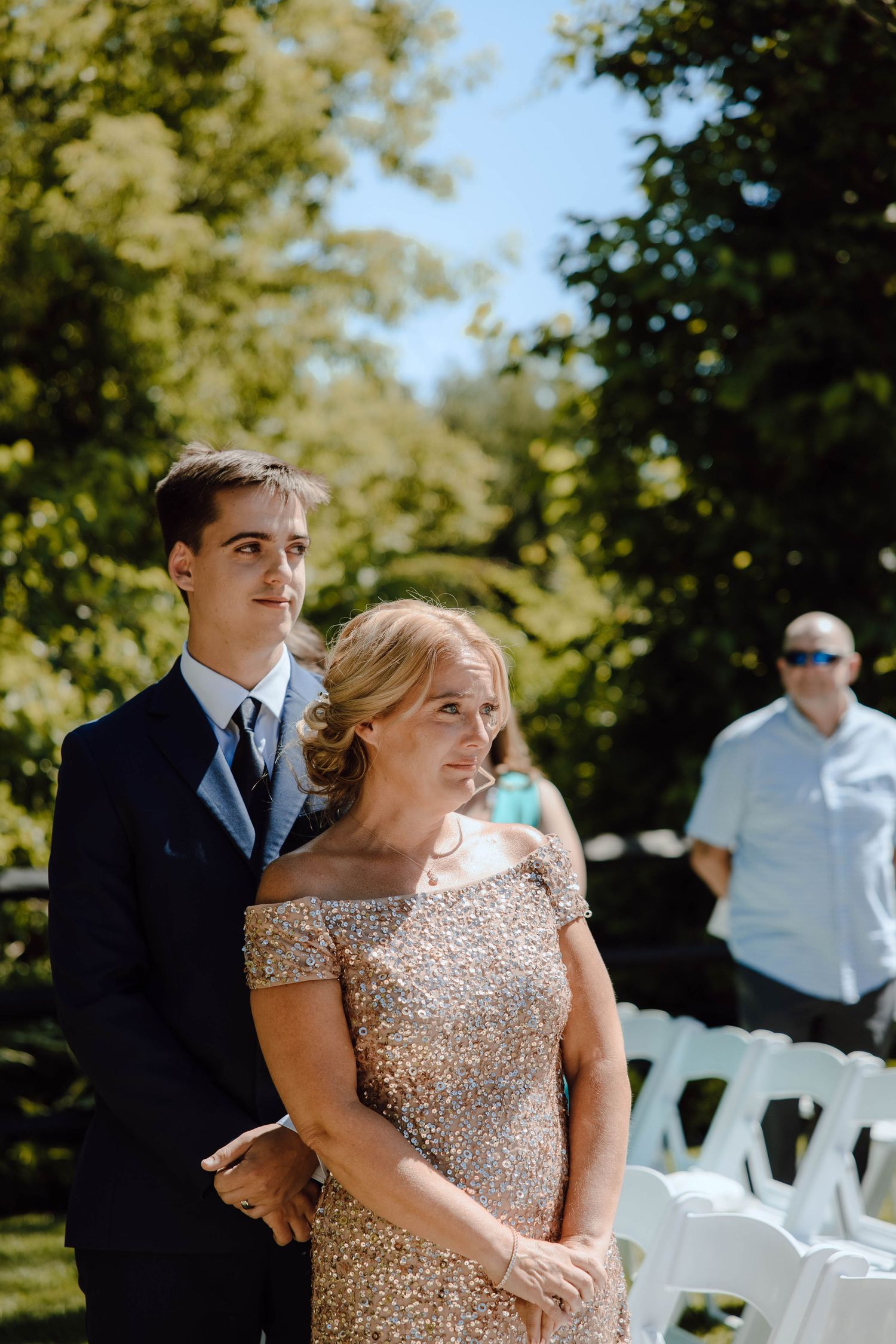 Young man in suit escorts elegantly dressed woman in gold sequin gown at outdoor wedding ceremony.