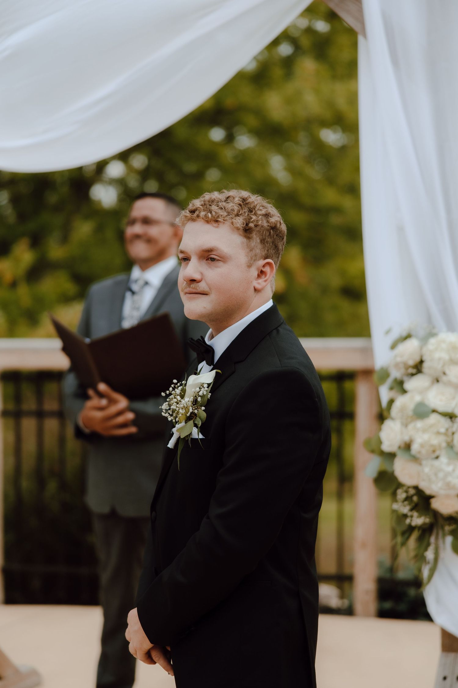 Groom in black tuxedo with floral boutonniere smiling at outdoor wedding ceremony with officiant in background.