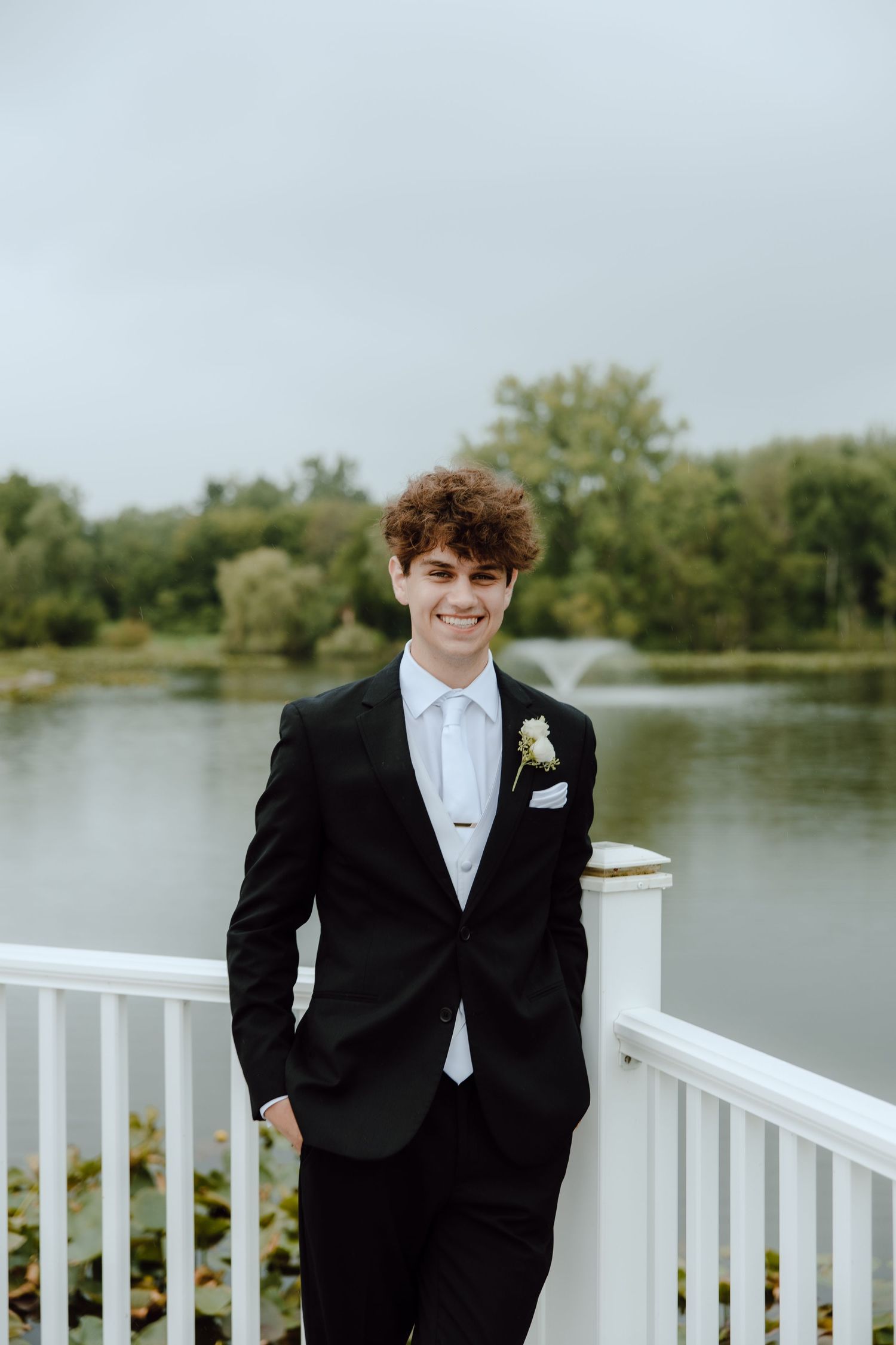 Smiling groom in black suit with boutonniere poses on white deck by pond.
