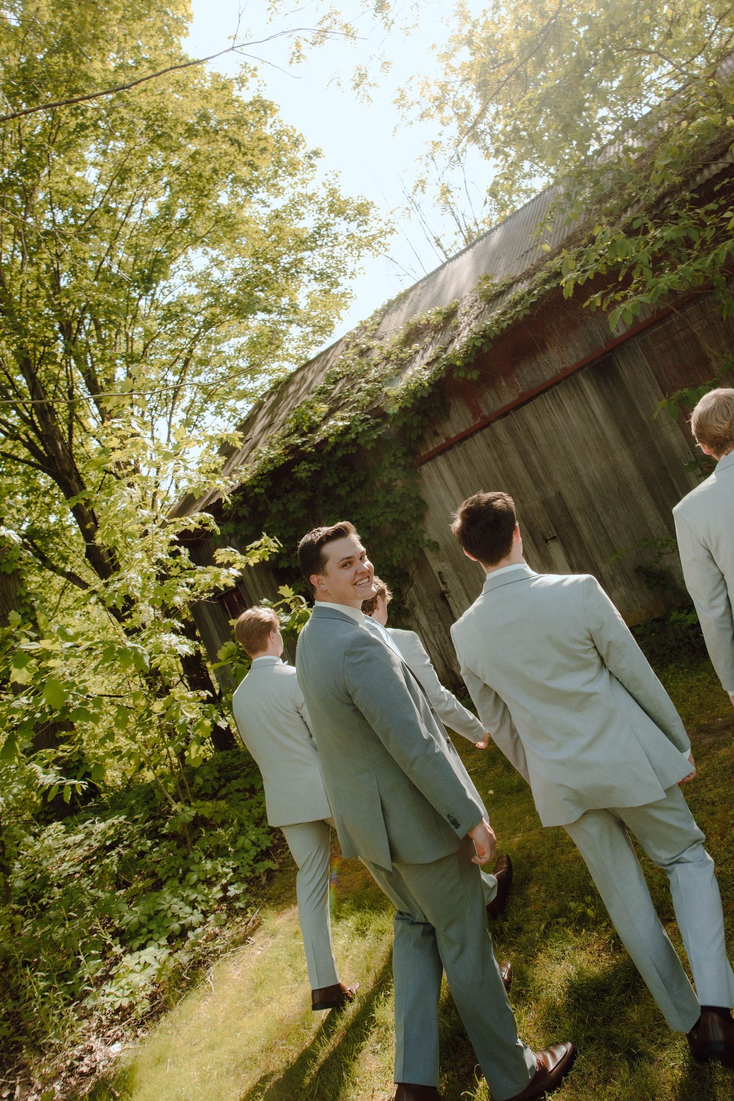 Groom smiles at camera with groomsmen in matching sage suits near a rustic barn surrounded by trees.