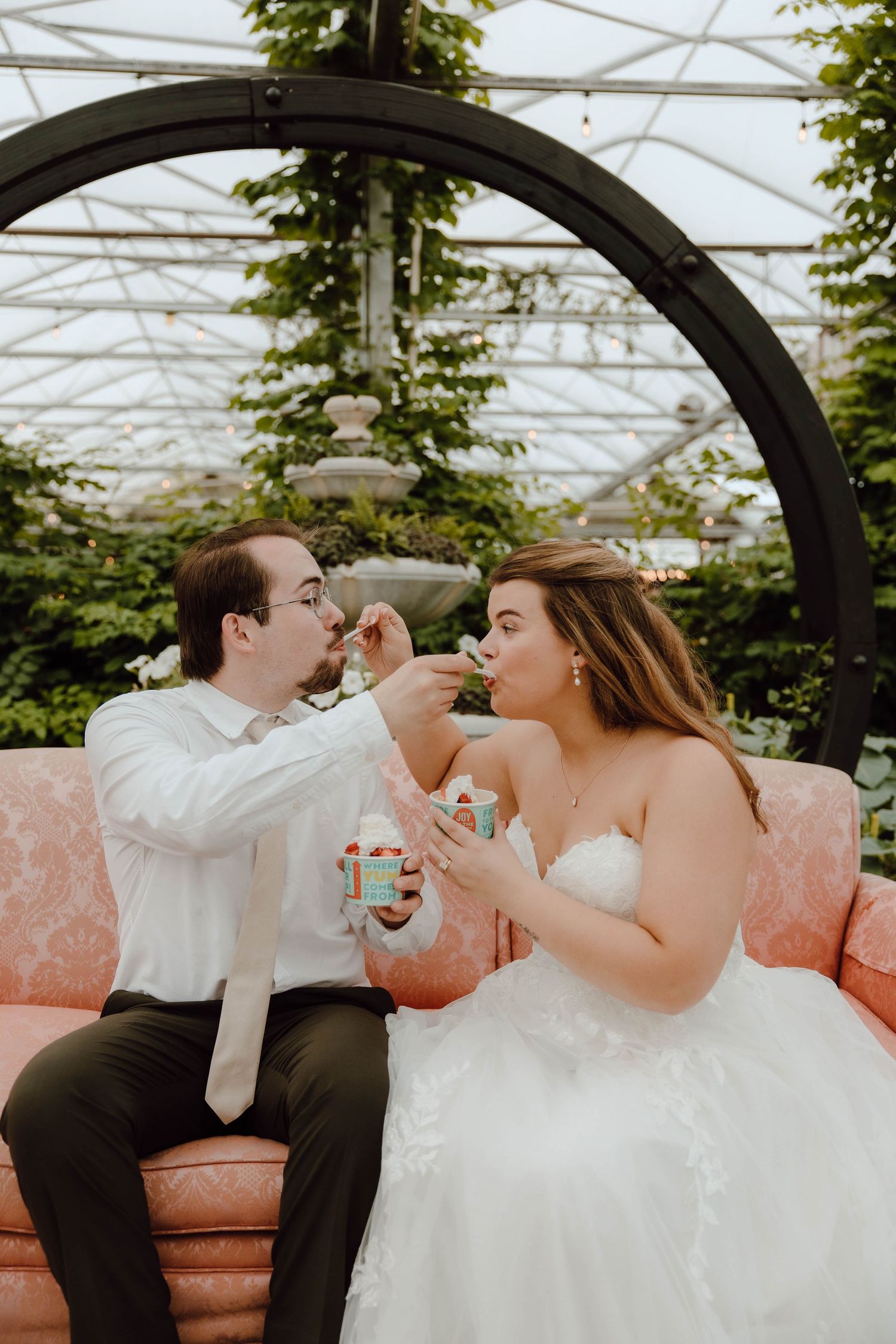 Bride and groom feed each other ice cream while seated on a pink sofa inside a greenhouse wedding venue.