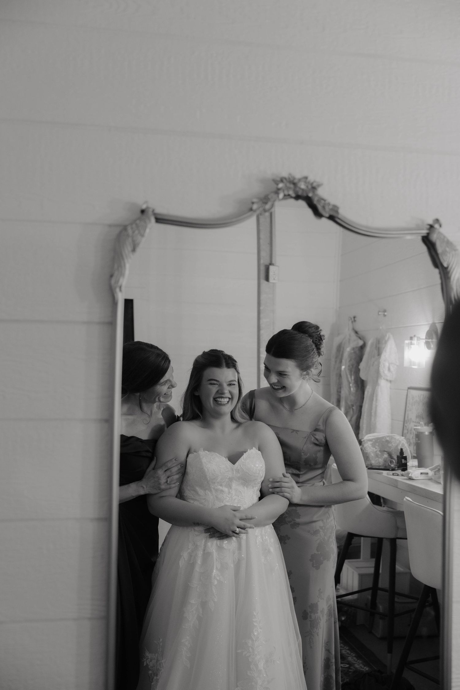 Bride laughing with bridesmaid in mirror reflection during wedding preparations in black and white photo.
