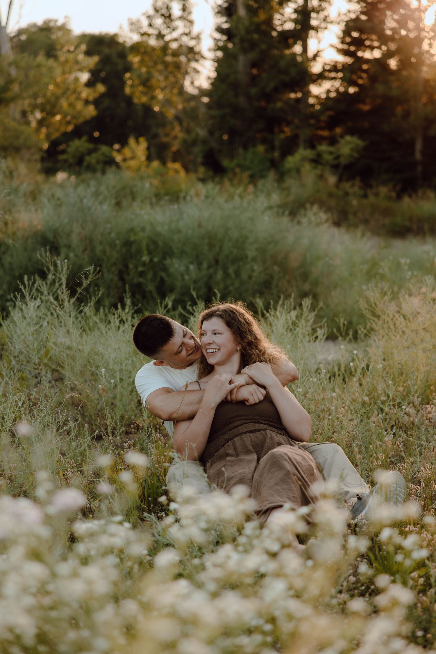 Couple sitting in wildflower field at golden hour, laughing and embracing romantically outdoors.