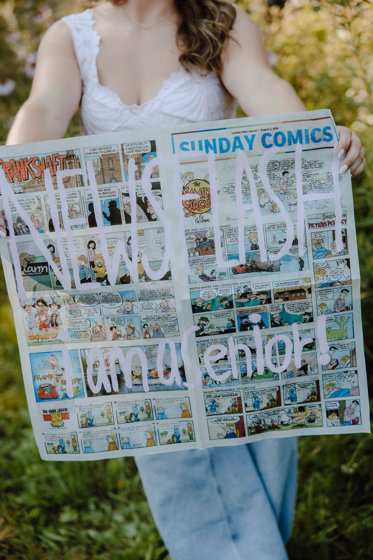 Bride holding Sunday Comics newspaper with handwritten wedding message outdoors in green garden setting.
