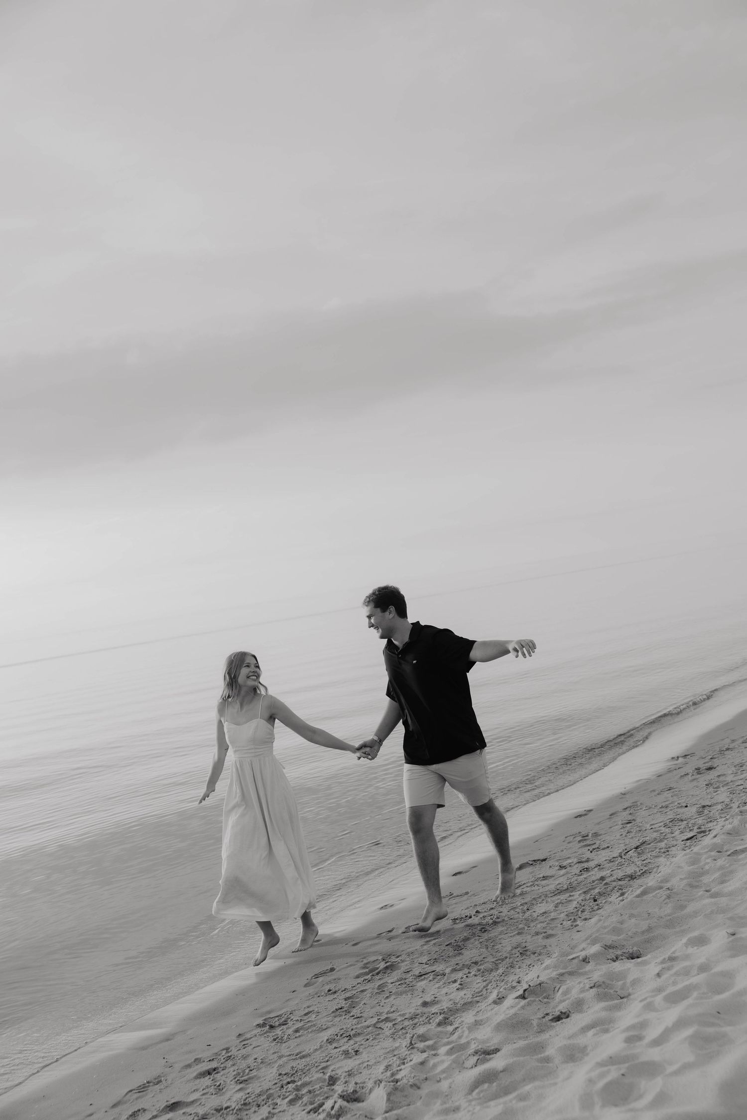 Black and white photo of a couple holding hands and walking barefoot along a sandy beach shore.
