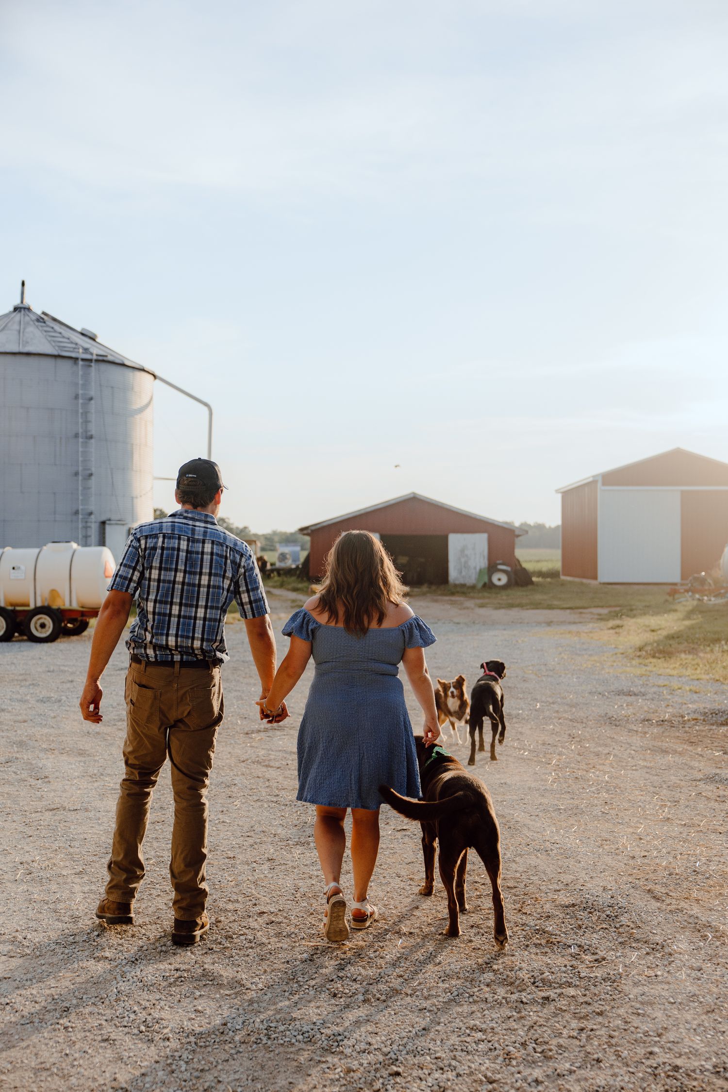 A couple walks with their dog on a farm path near grain silos at sunset.