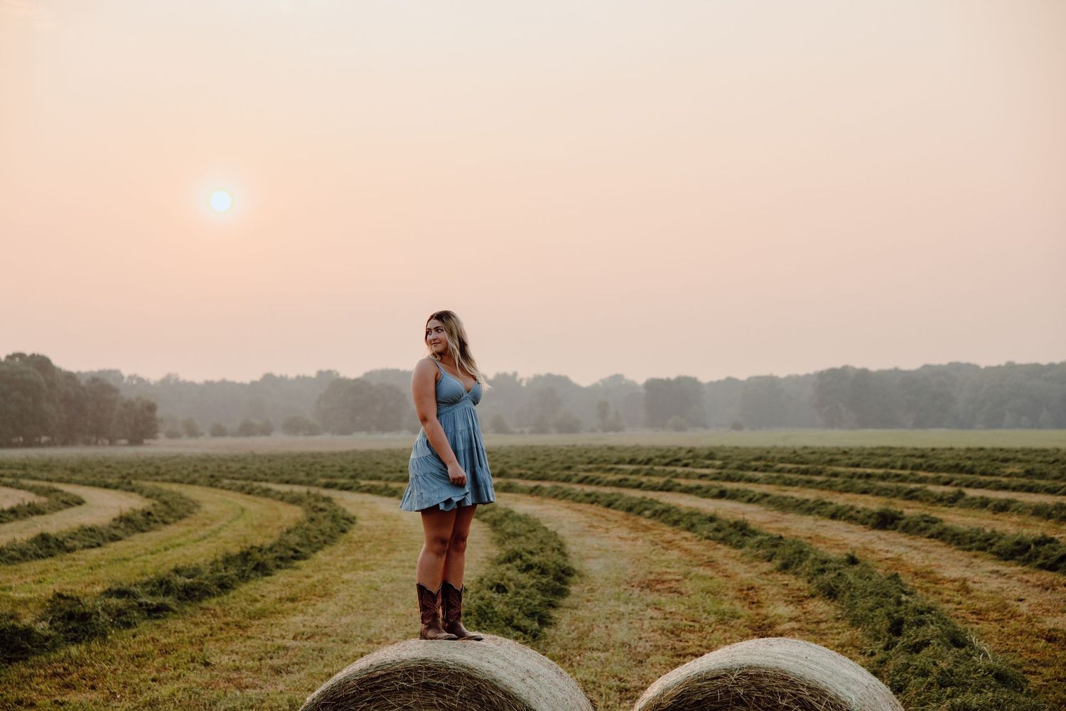 Two people pose playfully on hay bales in a golden sunset-lit field.