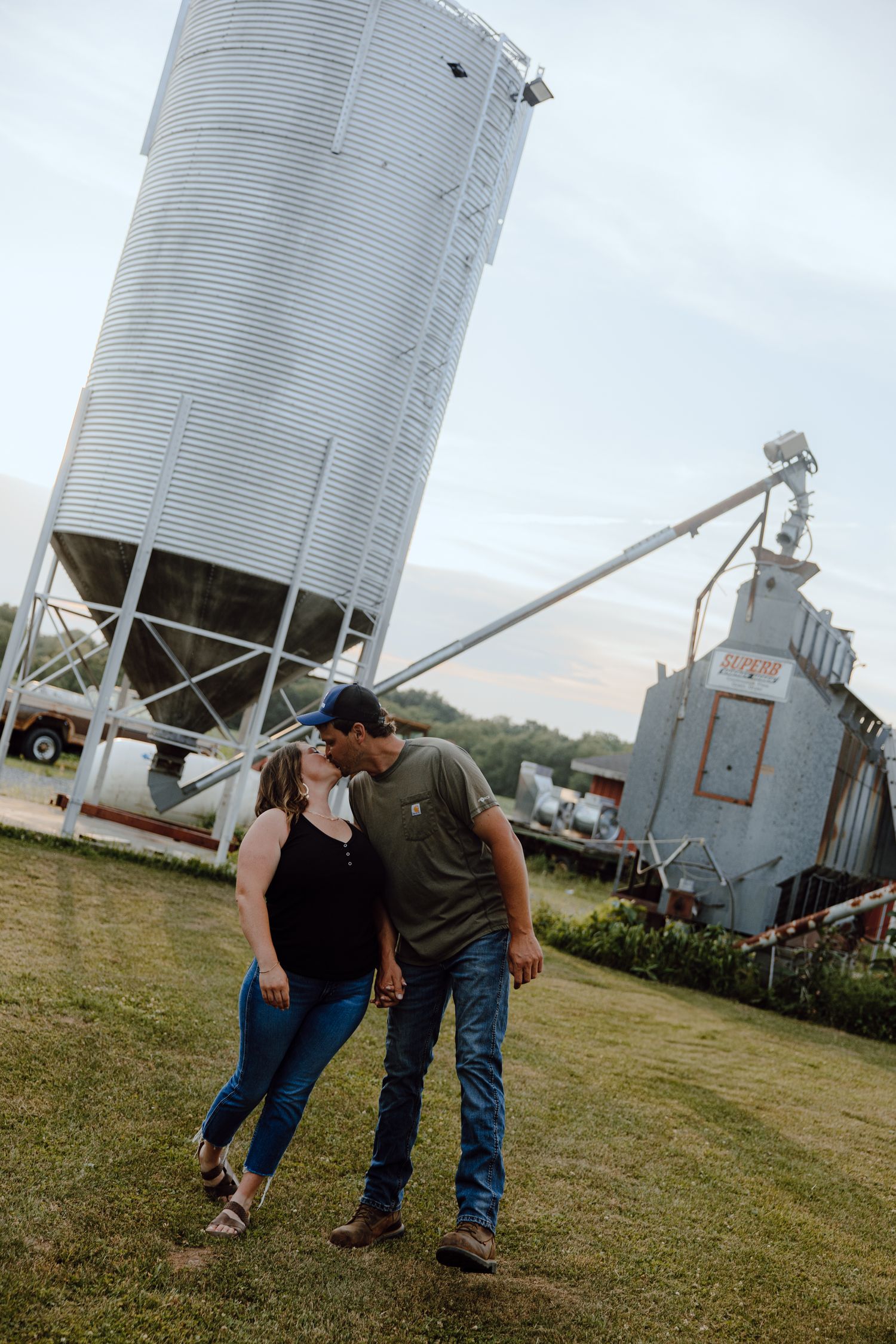 A romantic couple embraces near a large grain silo and farm equipment at sunset.