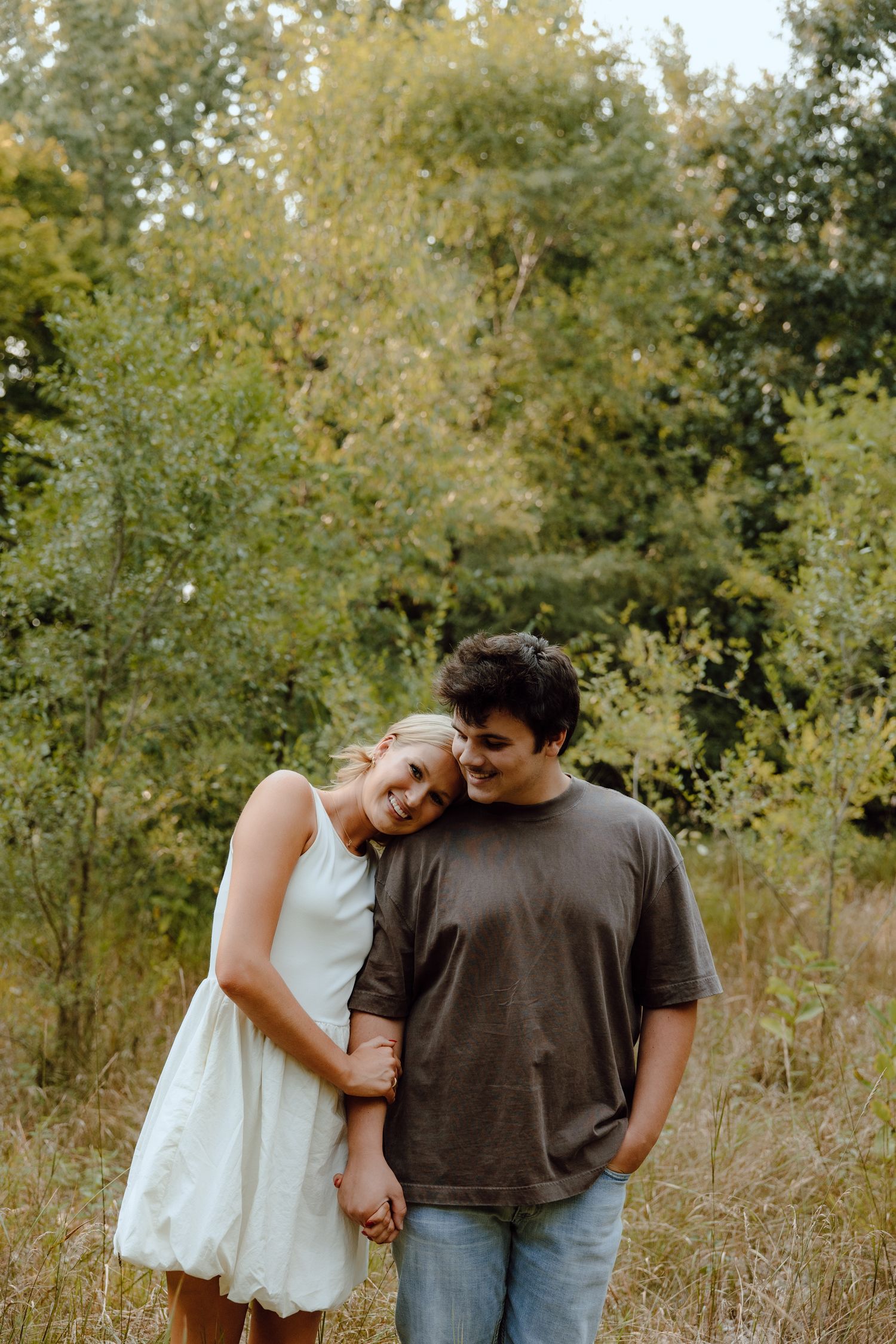 A couple shares an intimate moment while standing in tall grass and trees.