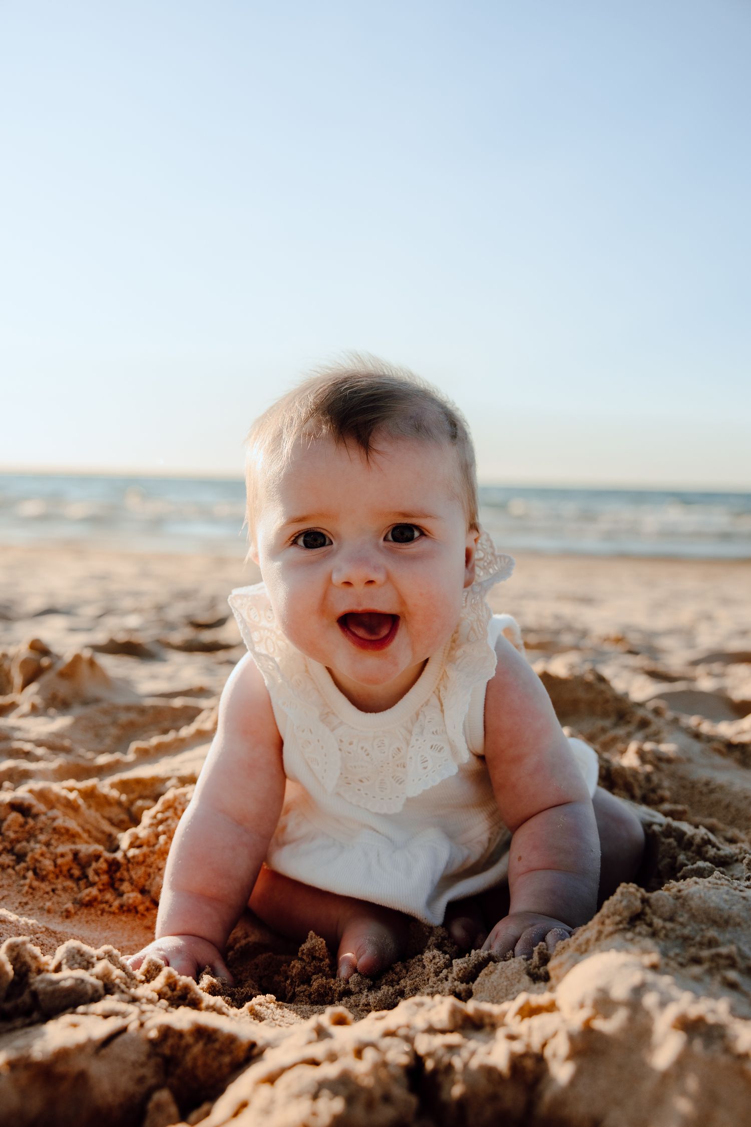 Baby in white outfit crawls happily on sandy beach with ocean waves in background.