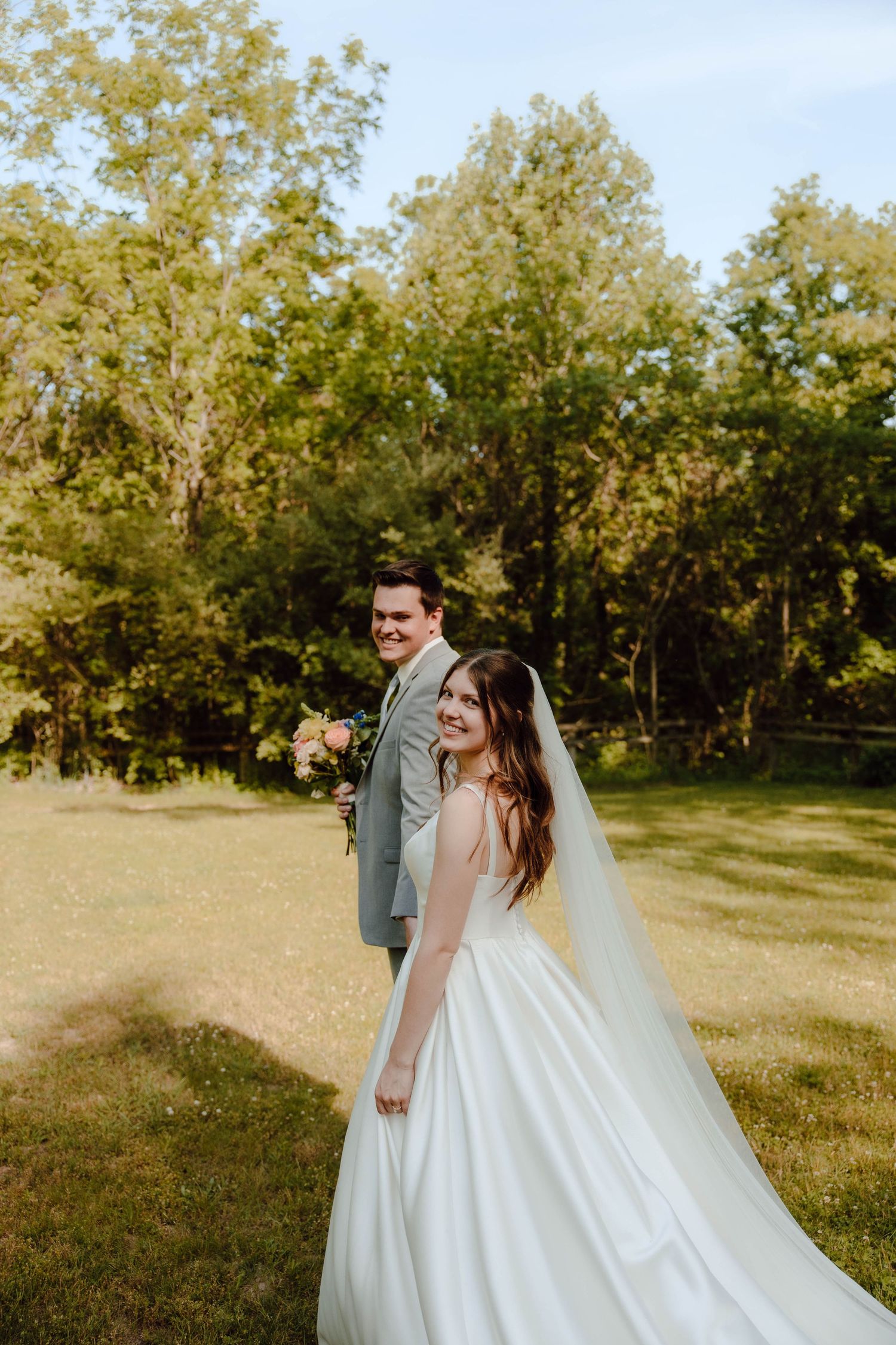 Bride in white ball gown and veil smiles over shoulder while groom in gray suit holds bouquet outdoors.