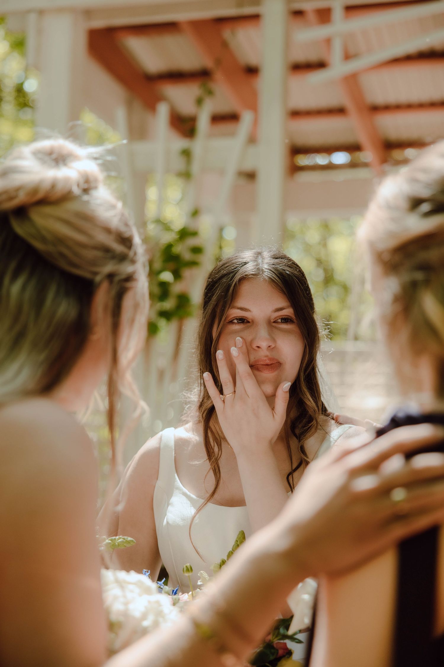 Emotional bride wiping tears at outdoor wedding ceremony surrounded by bridesmaids in white dress.