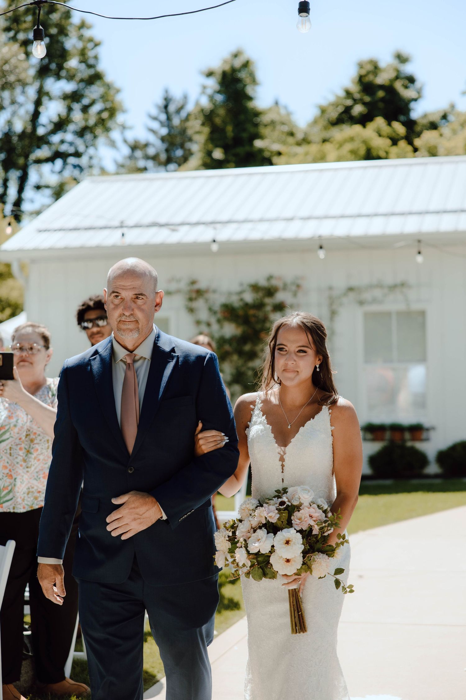 Bride in lace gown walks down outdoor aisle with escort, holding pink bouquet, white barn venue in background.