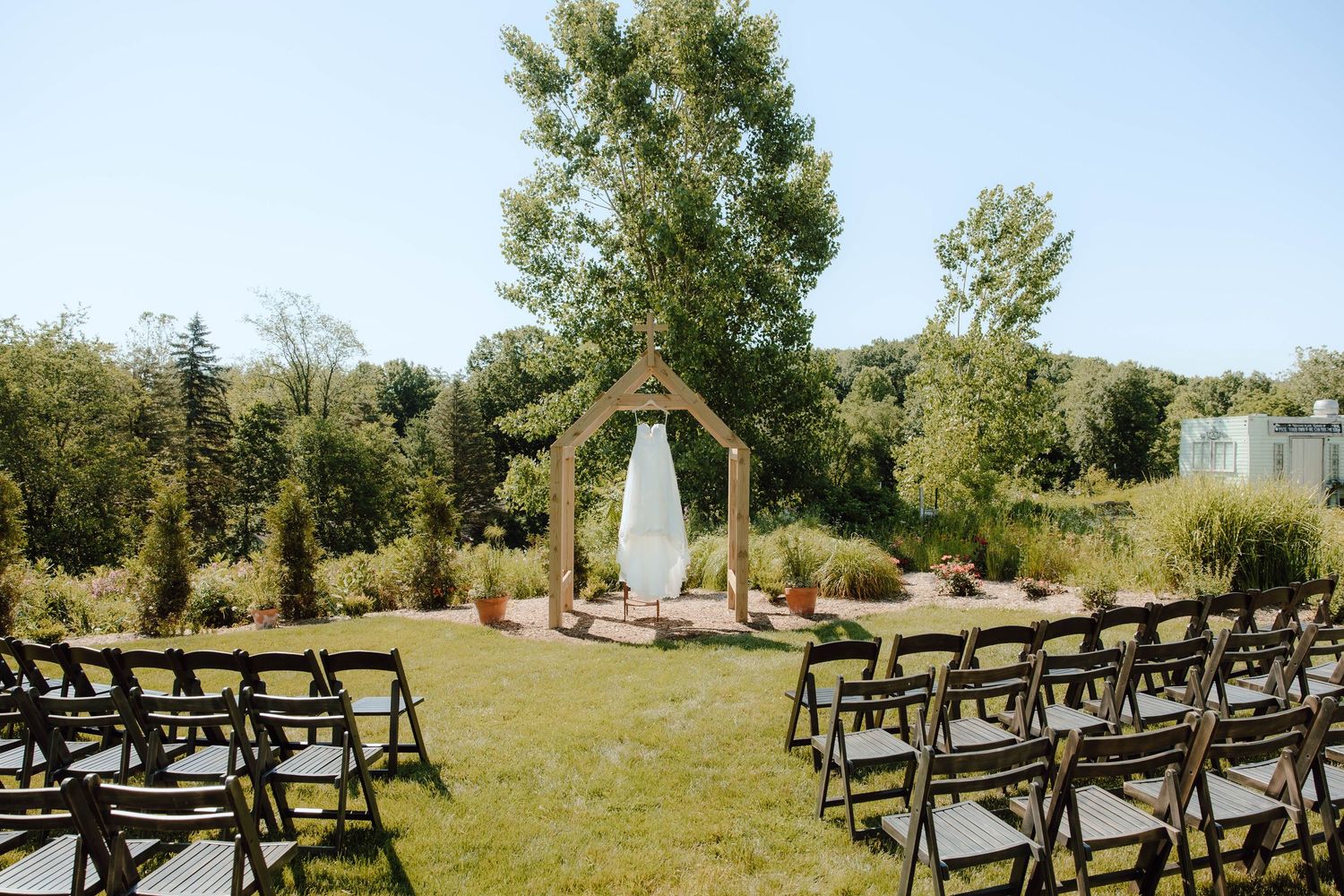 Outdoor wedding ceremony setup with wooden arch, folding chairs on green lawn, surrounded by lush gardens on a sunny day.