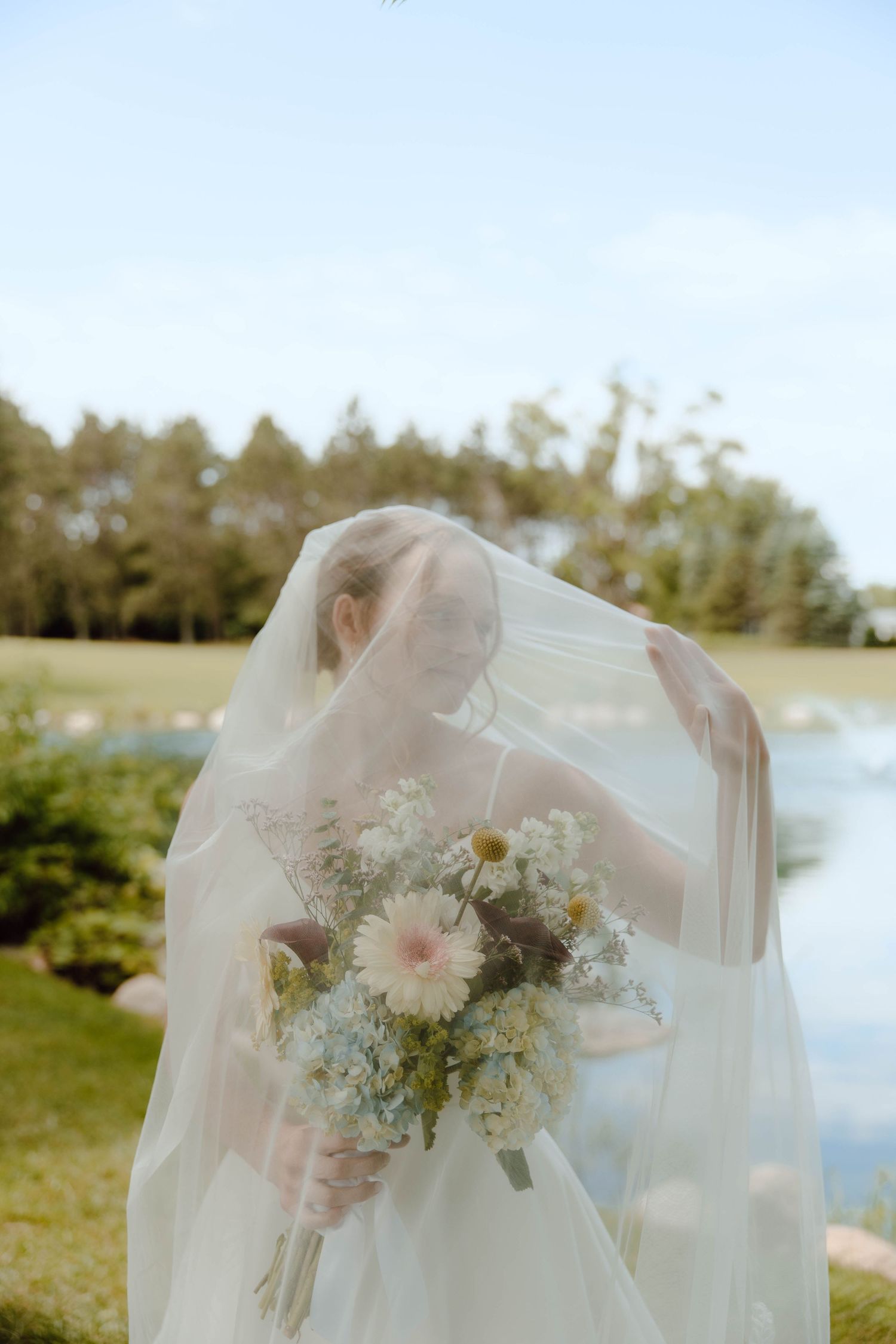 Bride in white wedding dress and veil holding a soft pastel bouquet outdoors near a serene lake surrounded by trees.
