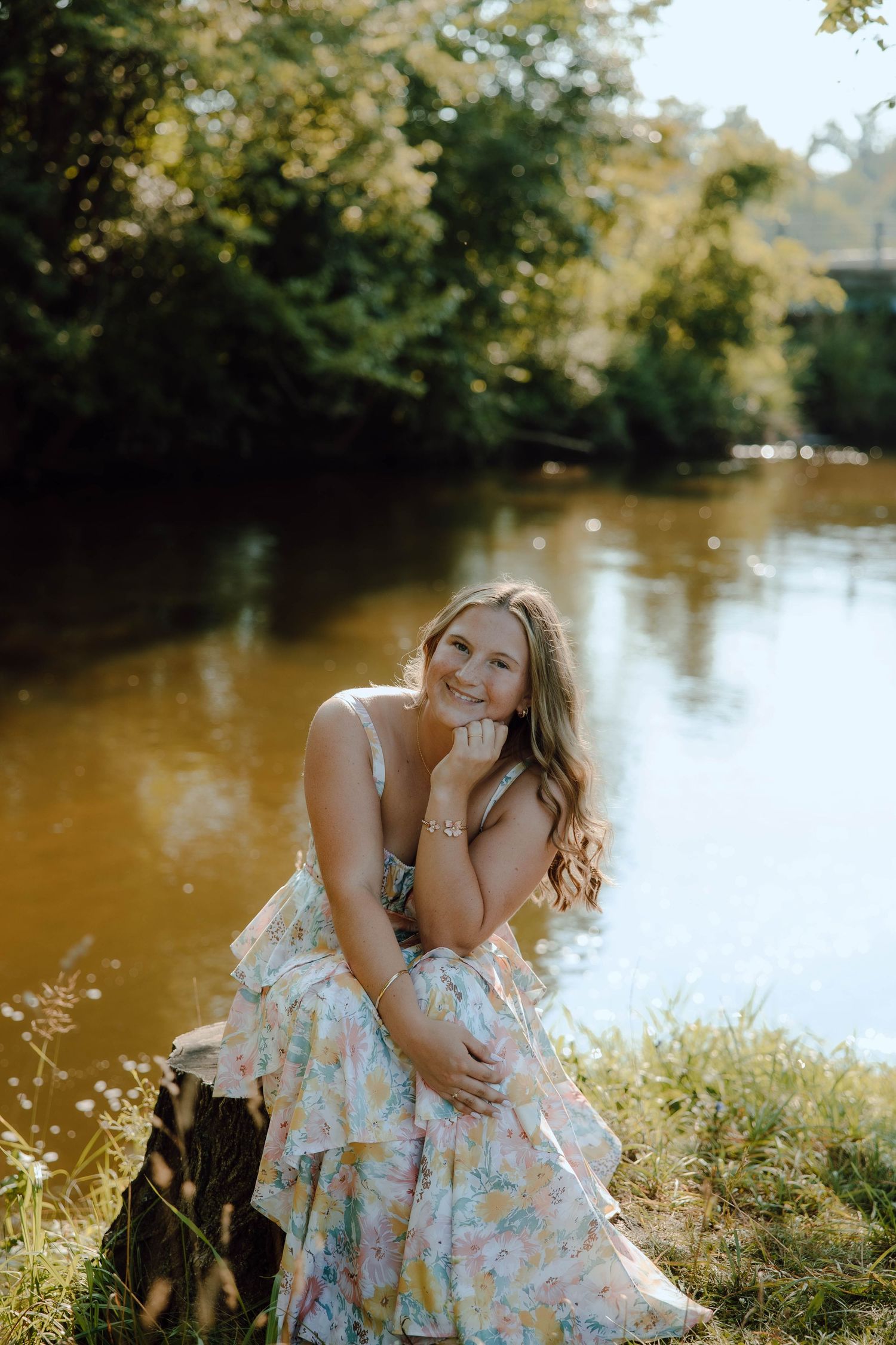 Young woman in floral dress sitting by a sunlit river, smiling warmly, surrounded by lush green trees.