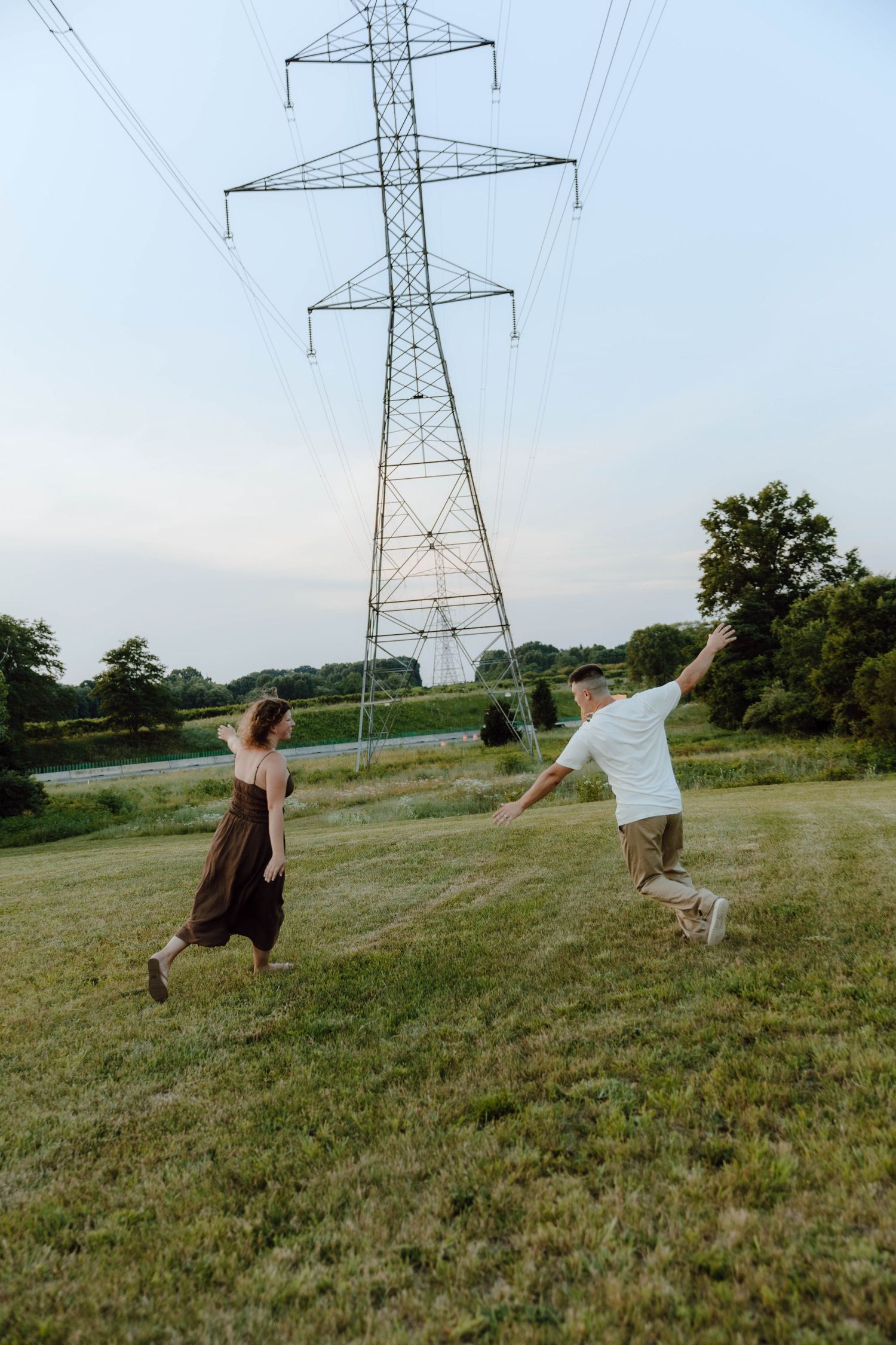 Couple playfully running in a green field near a large electrical transmission tower on a cloudy day.
