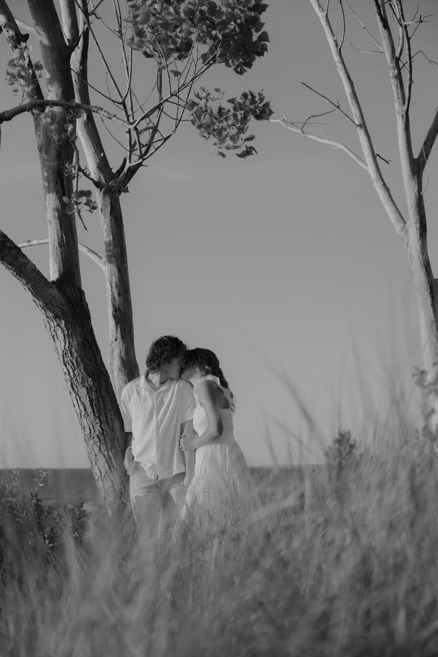 Black and white photo of a romantic couple embracing among bare trees in a grassy field.