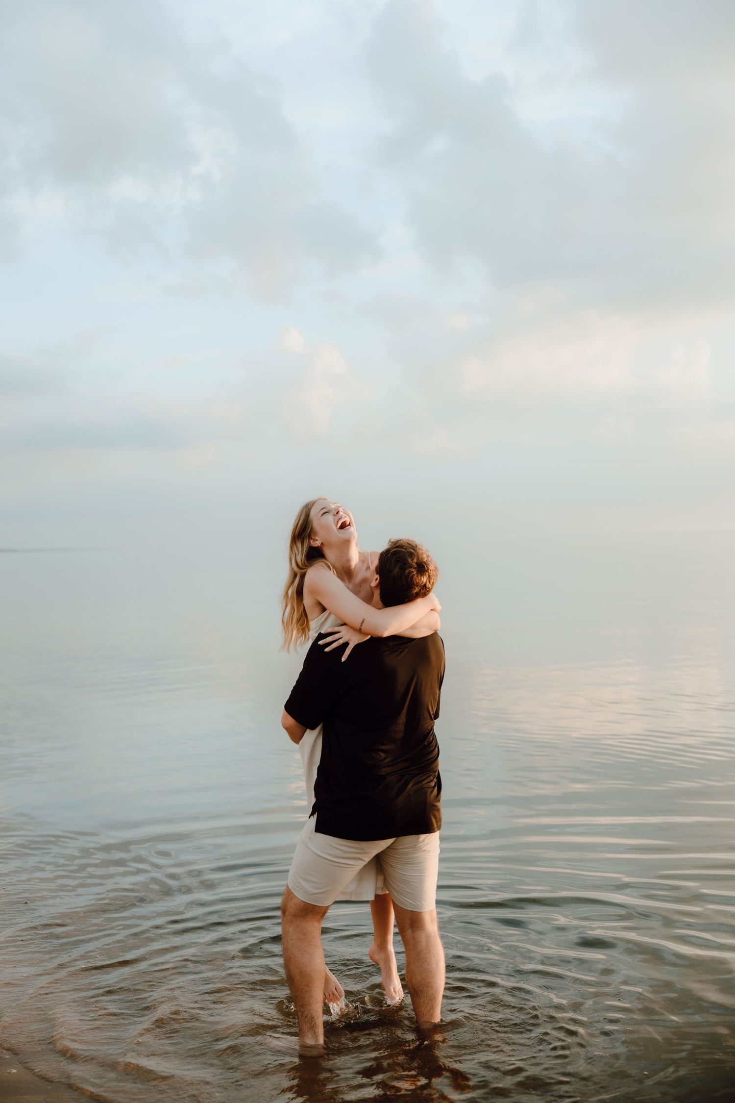 A couple laughing and embracing while standing in shallow ocean water at dusk on a cloudy beach.