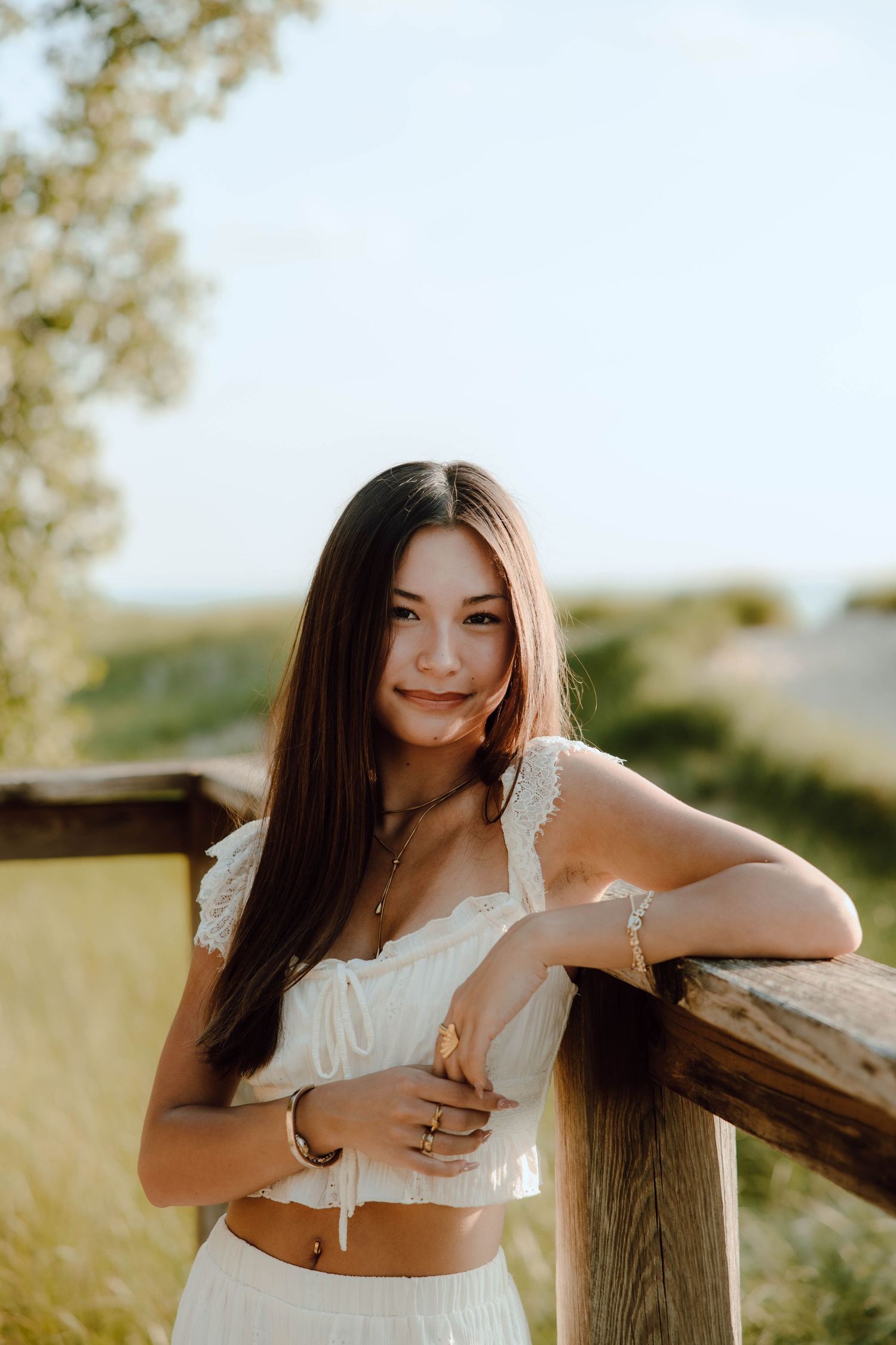 Young woman in white crop top leaning on wooden fence in sunny field, smiling softly with long dark hair.