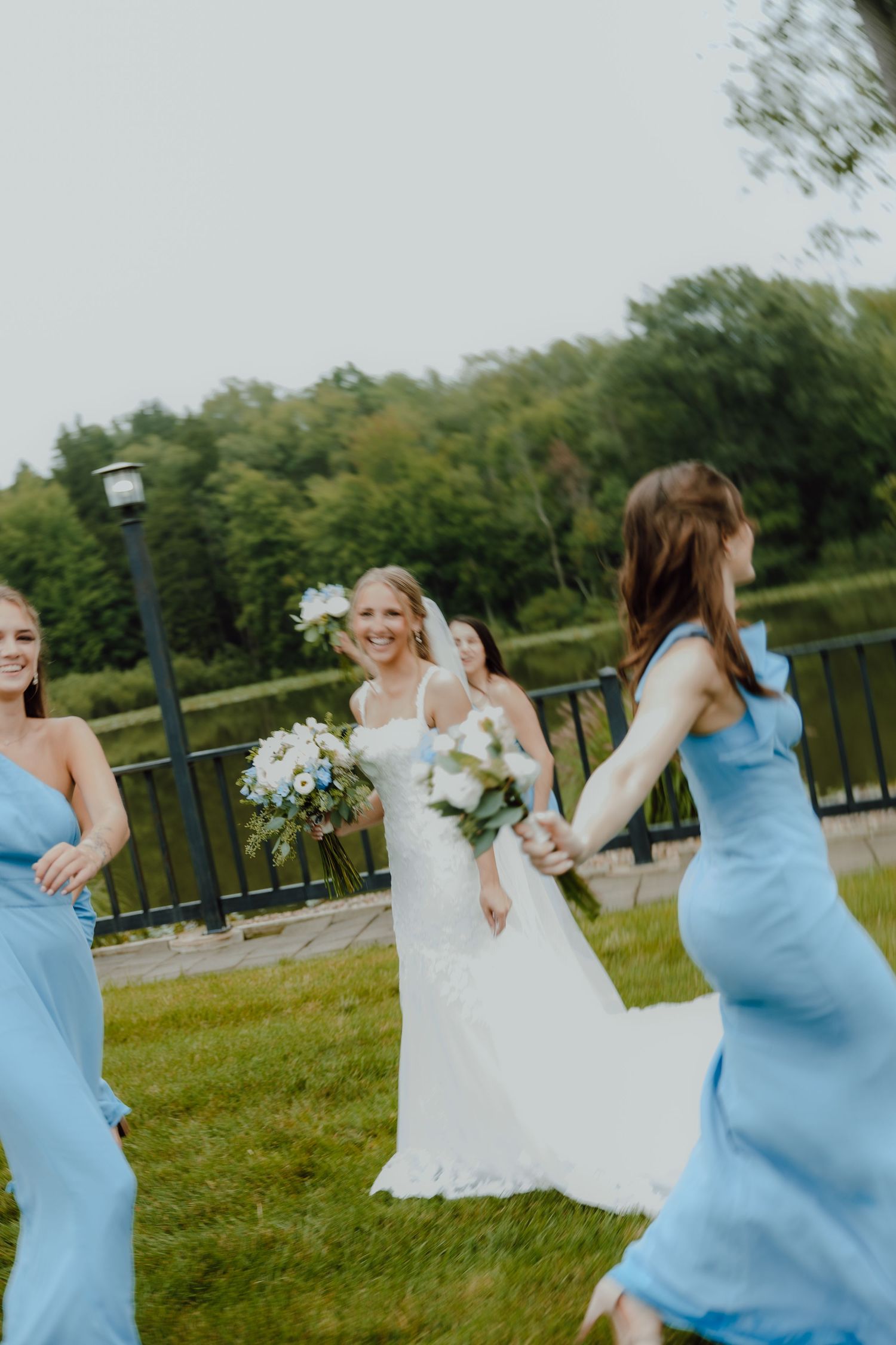 Bride in white lace gown walks with bridesmaids in light blue dresses, holding bouquets outdoors.