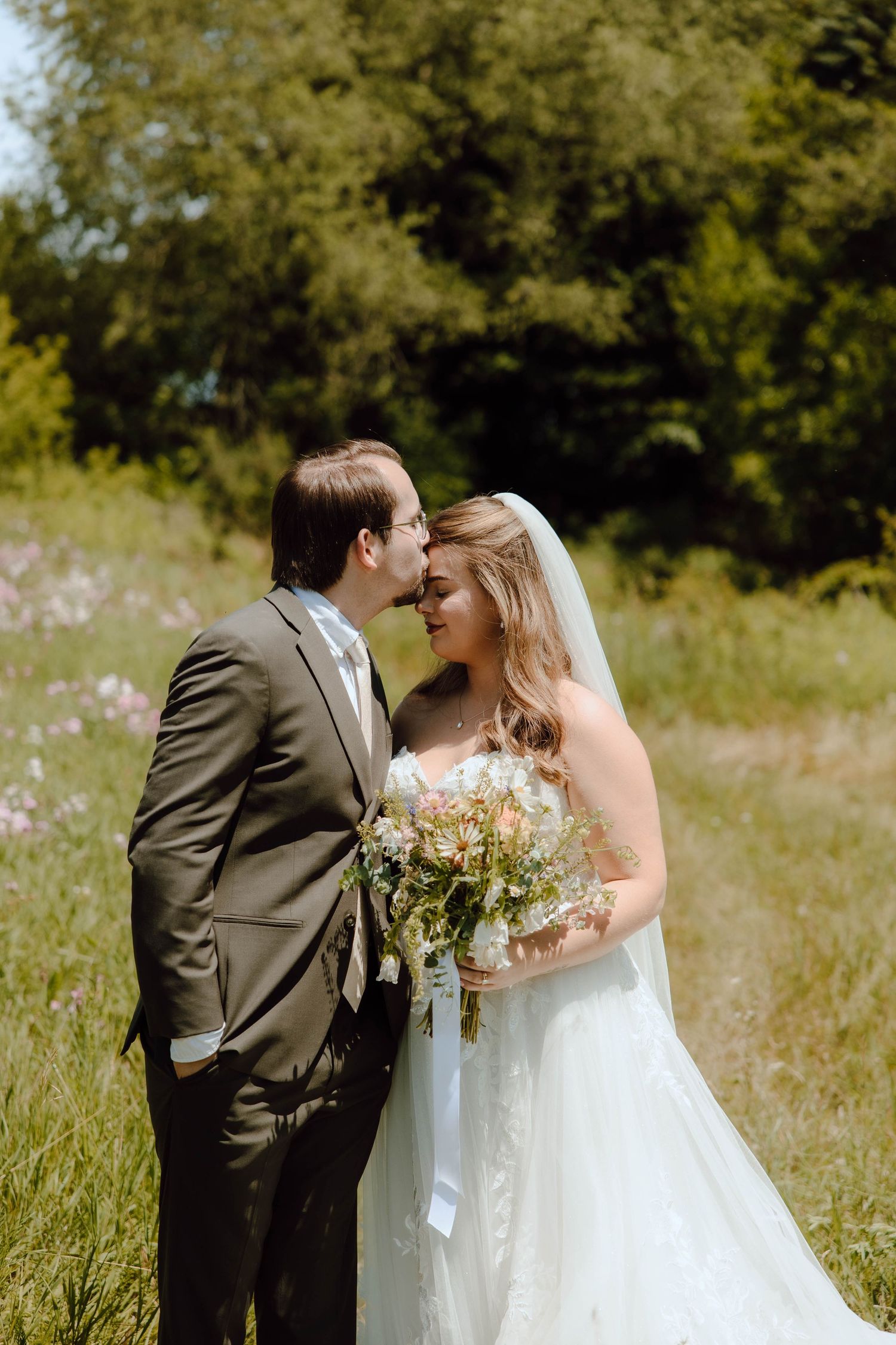 Bride and groom share an intimate moment in a sunlit meadow, bride holding a wildflower bouquet in a lace gown.