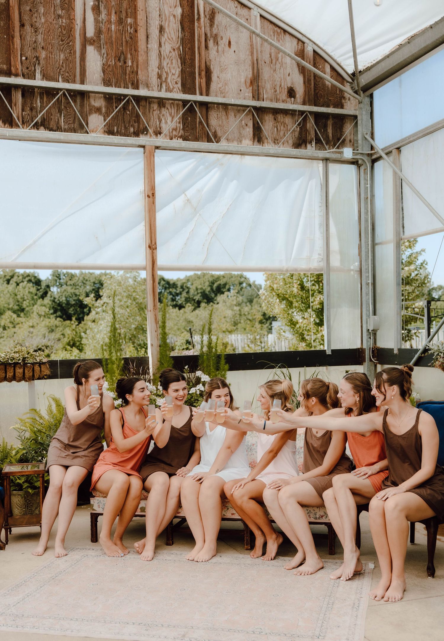 Bridesmaids in earth-tone dresses toast together inside a rustic greenhouse venue before the wedding.