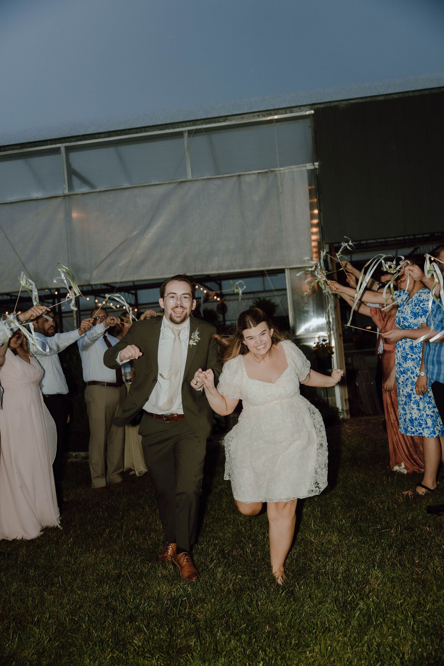 Newlyweds joyfully run through a ribbon exit surrounded by cheering guests at evening reception.
