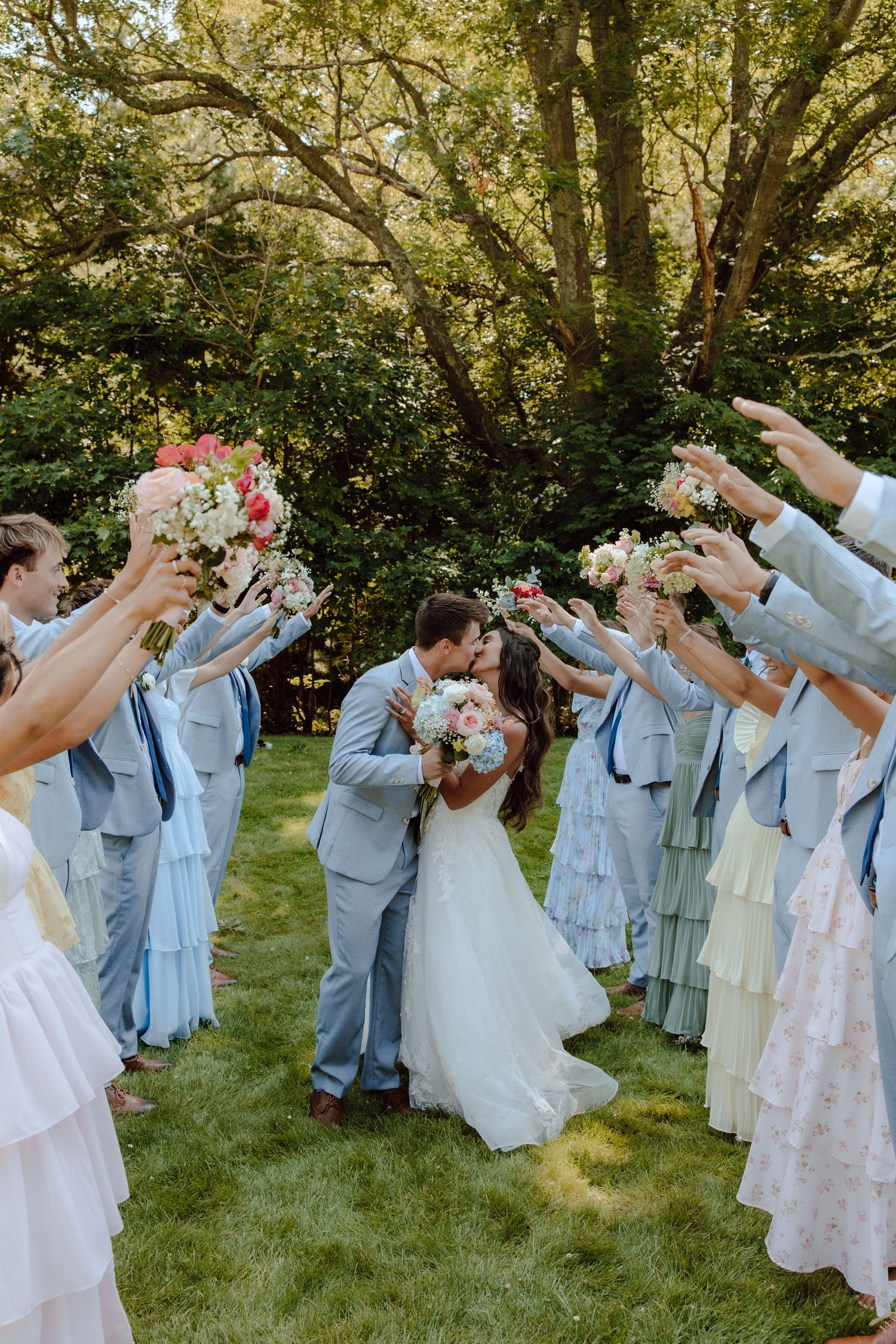 Wedding party raises bouquets as bride and groom share a kiss under floral arch on green lawn.