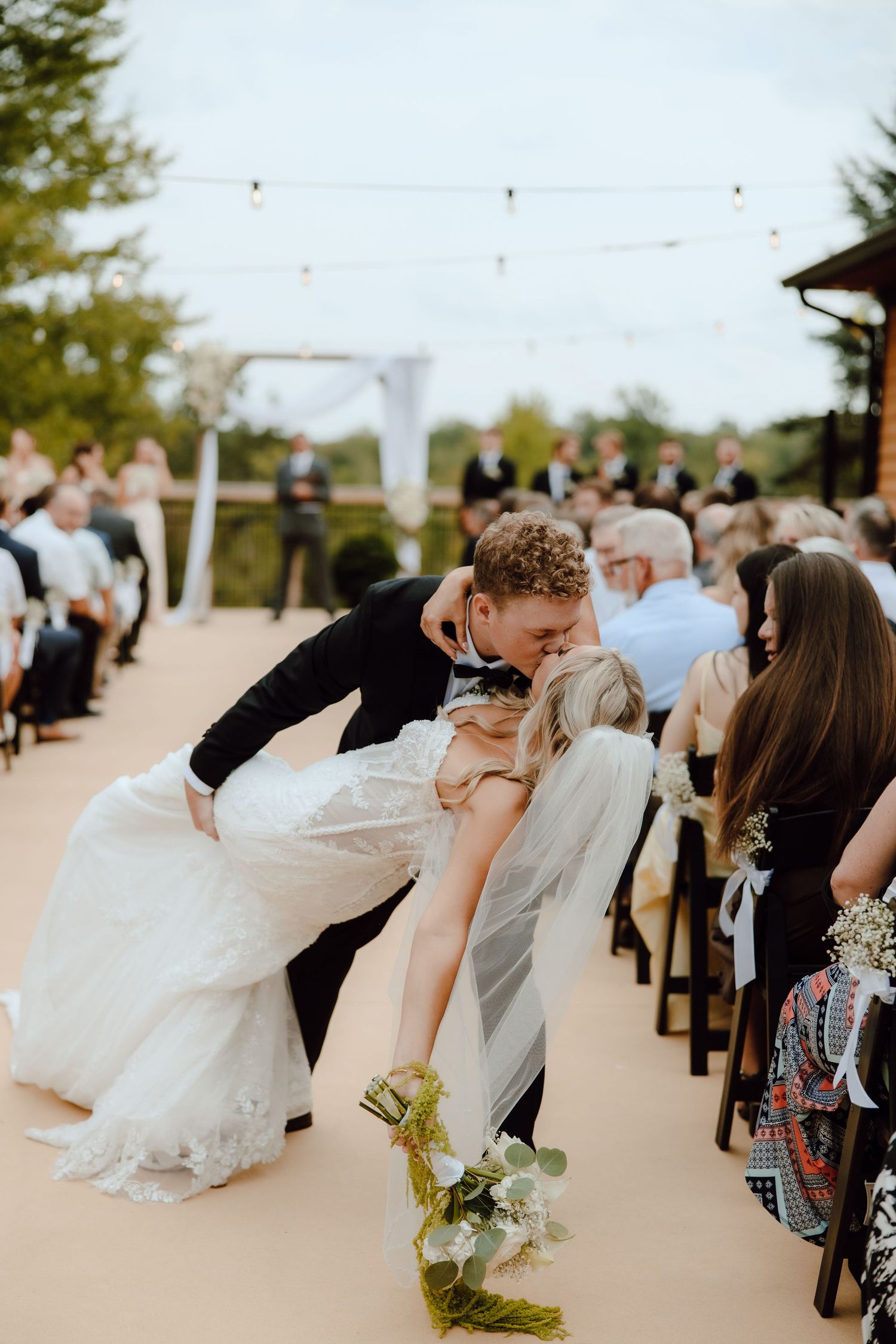 Groom dipping bride for a kiss during outdoor wedding ceremony with guests seated in rows.