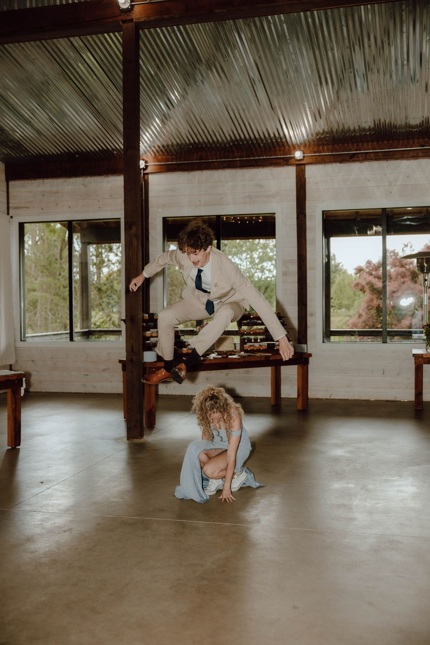 A groom in a suit leaps off a table over a crouching bride in a blue dress inside a rustic wedding venue.