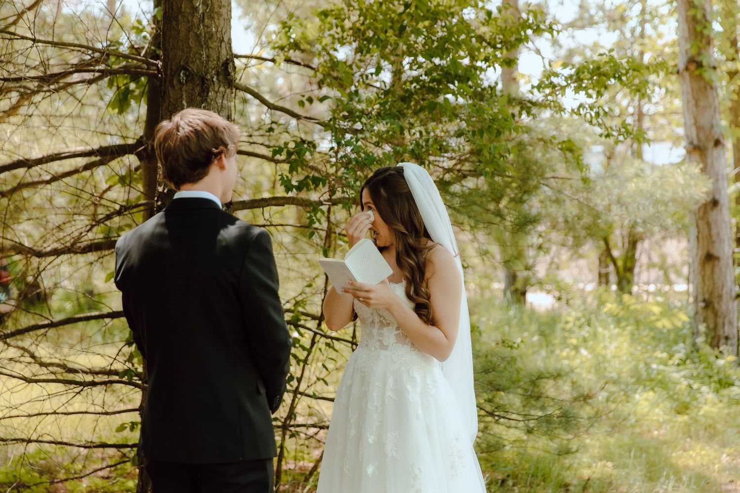 Bride reads wedding vows to groom in a lush forest setting, wearing a white gown and veil.