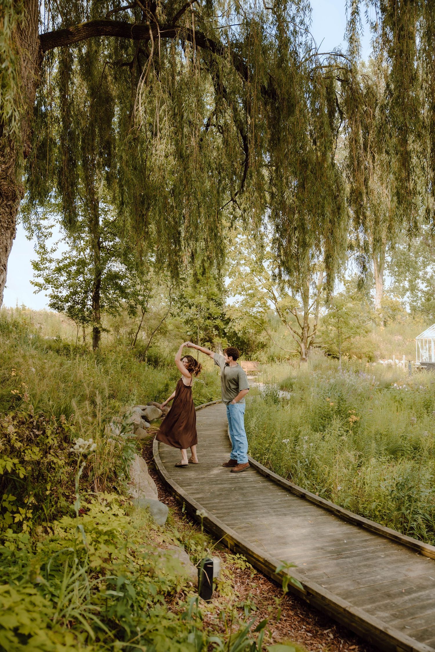 Couple dancing on a wooden boardwalk path surrounded by lush greenery and a weeping willow tree.