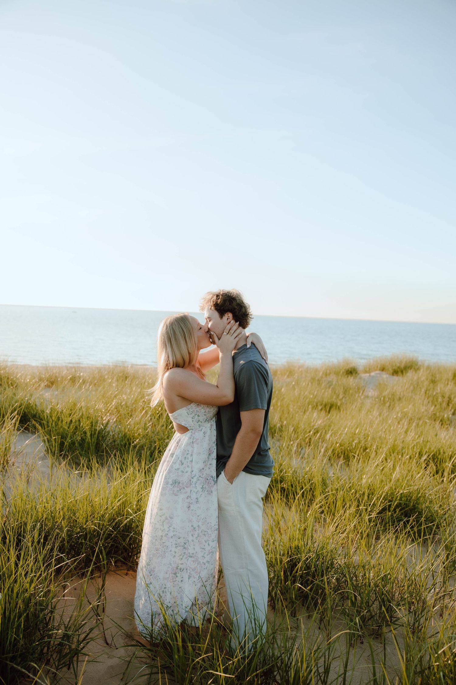 Couple kissing on sandy beach dunes with tall green grass and ocean in the background on a sunny day.